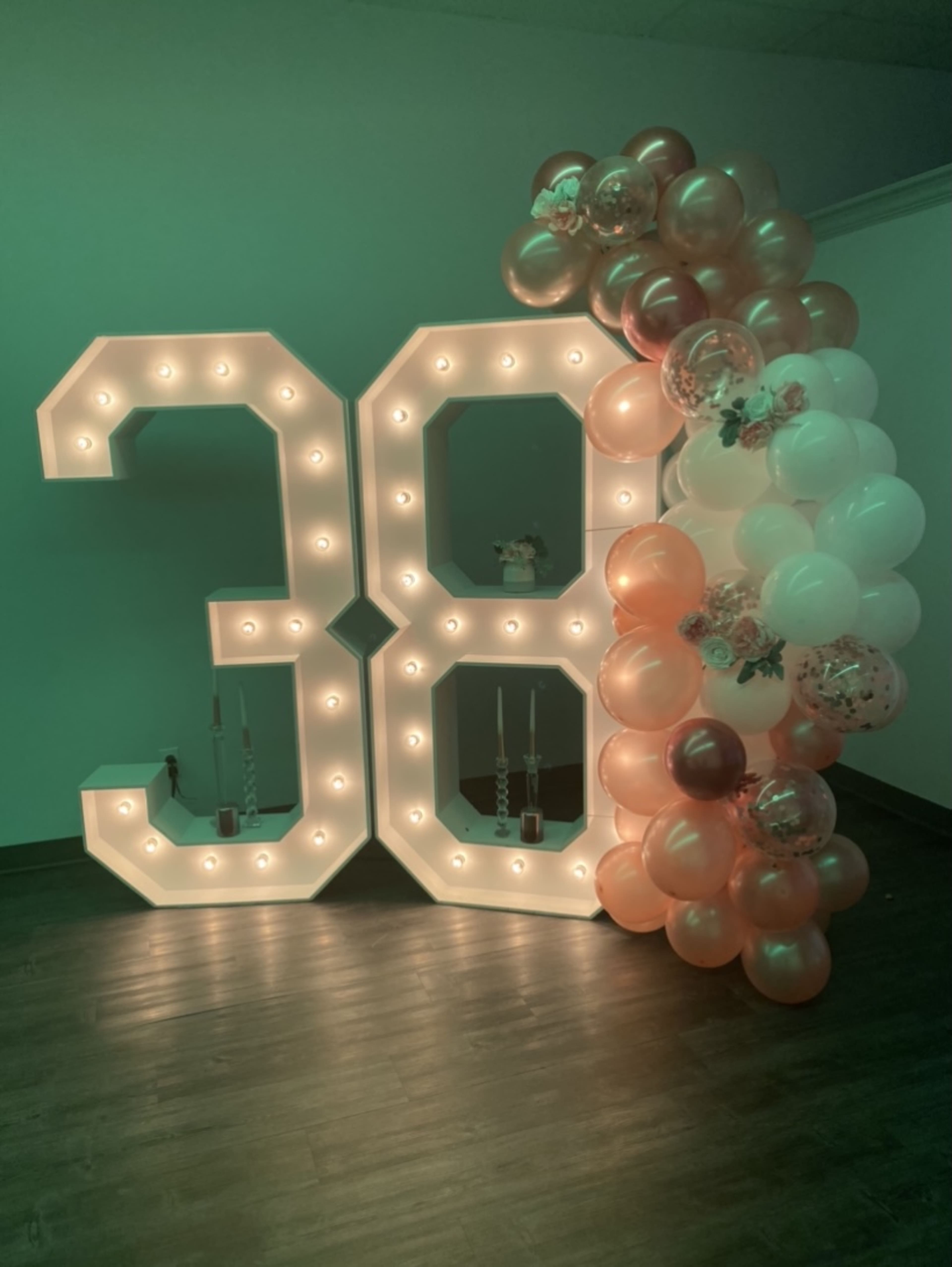 Large illuminated numbers "38" next to a decorative balloon arch in soft pink and white hues.
