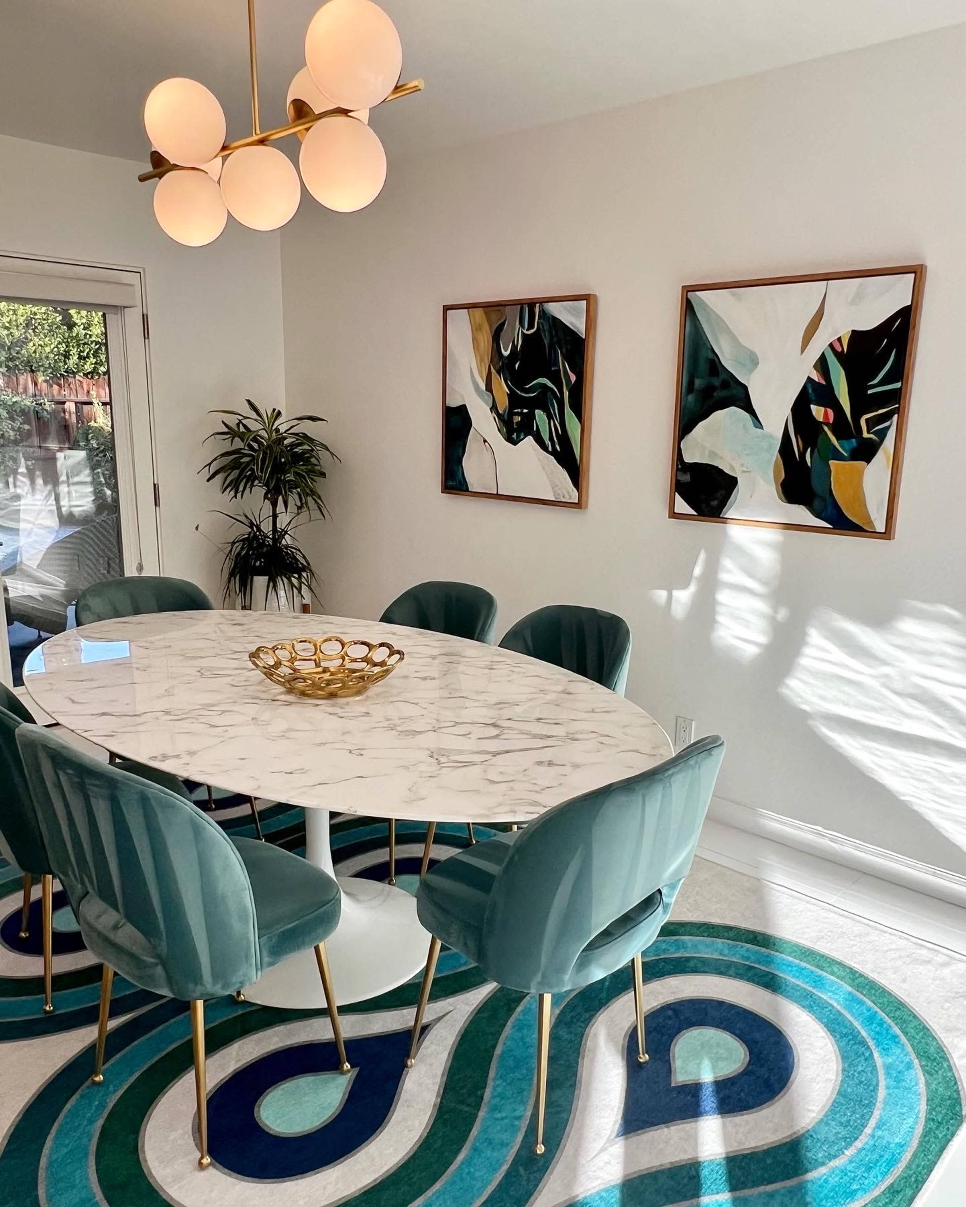 A modern dining area features a marble oval table surrounded by green velvet chairs, with a patterned rug and two framed artworks on the walls.