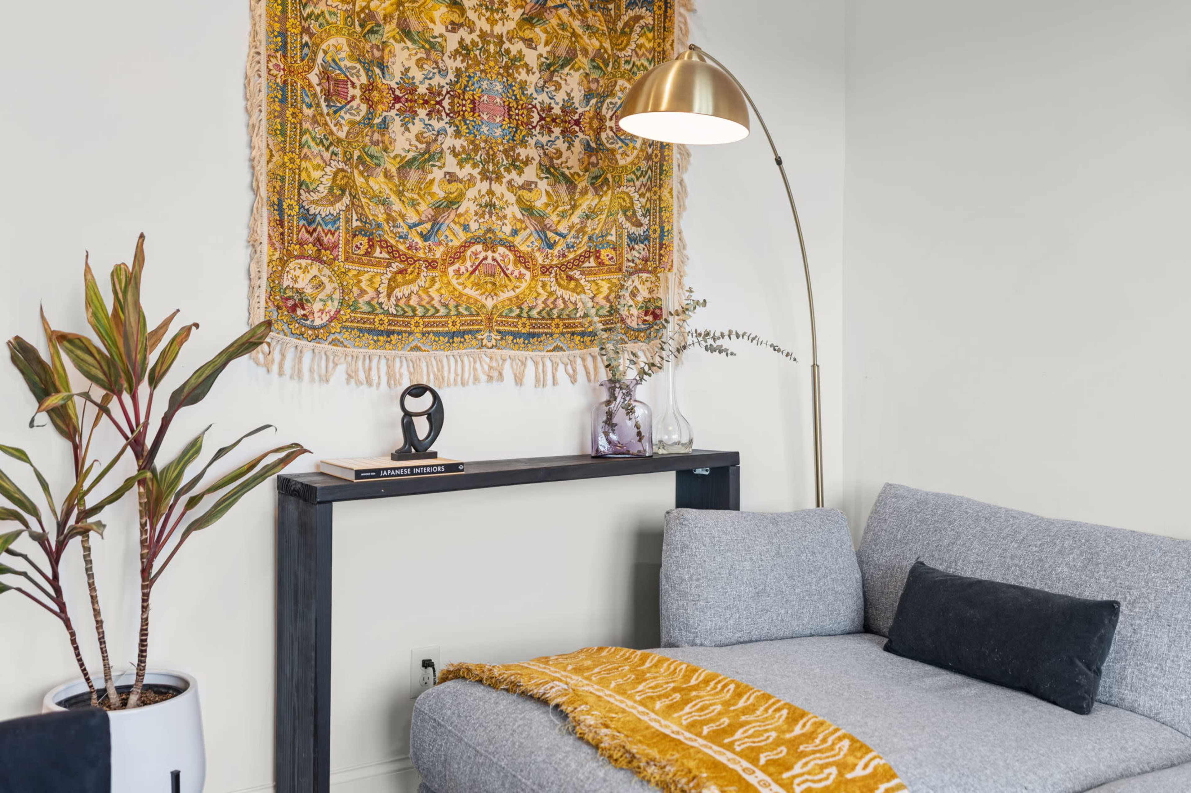 A cozy living space features a gray sectional sofa, a gold floor lamp, and a decorative wall tapestry above a console table with a plant and accessories.