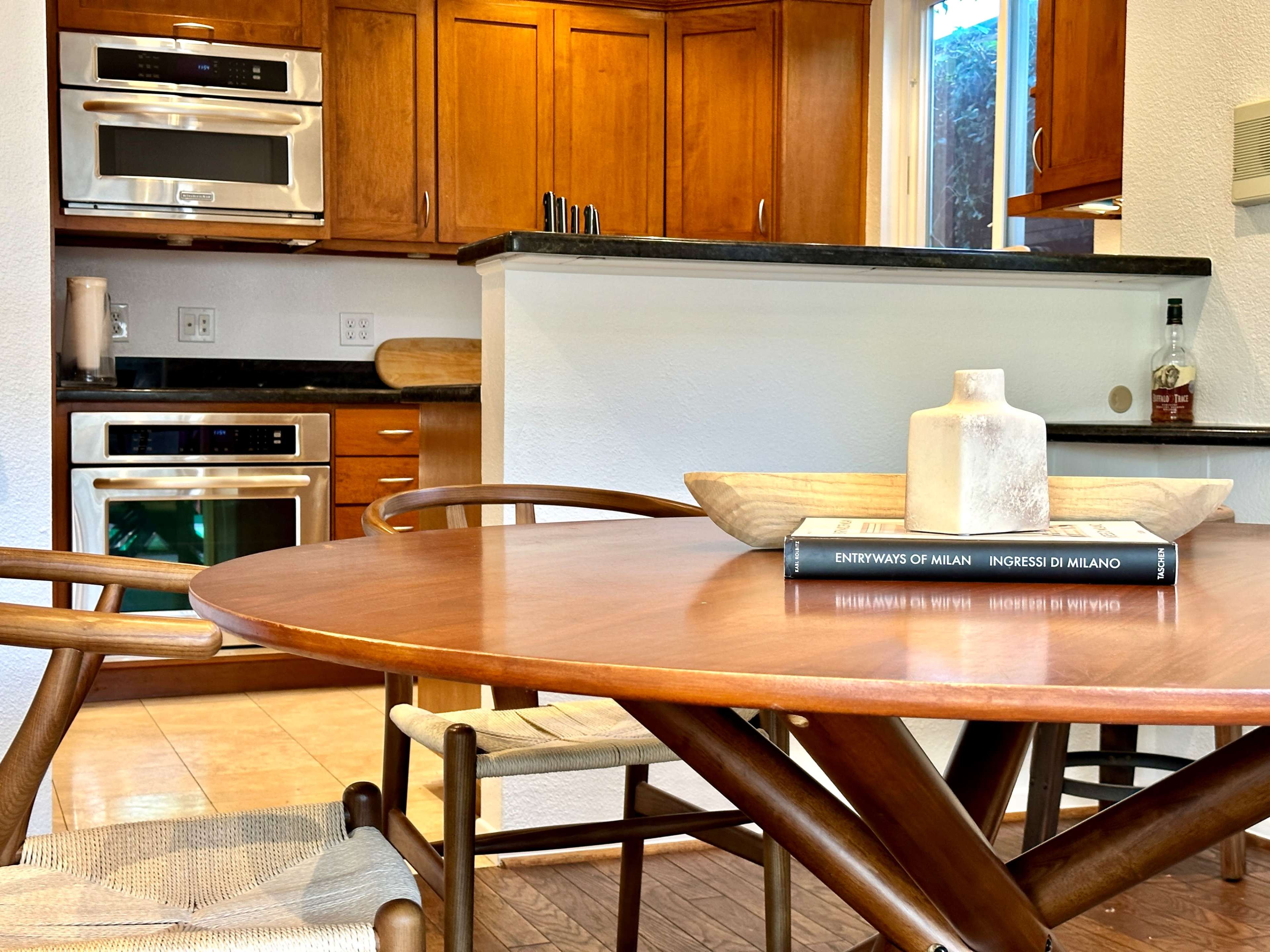 A wooden dining table with a decorative centerpiece is set in a modern kitchen featuring wooden cabinetry and stainless steel appliances.