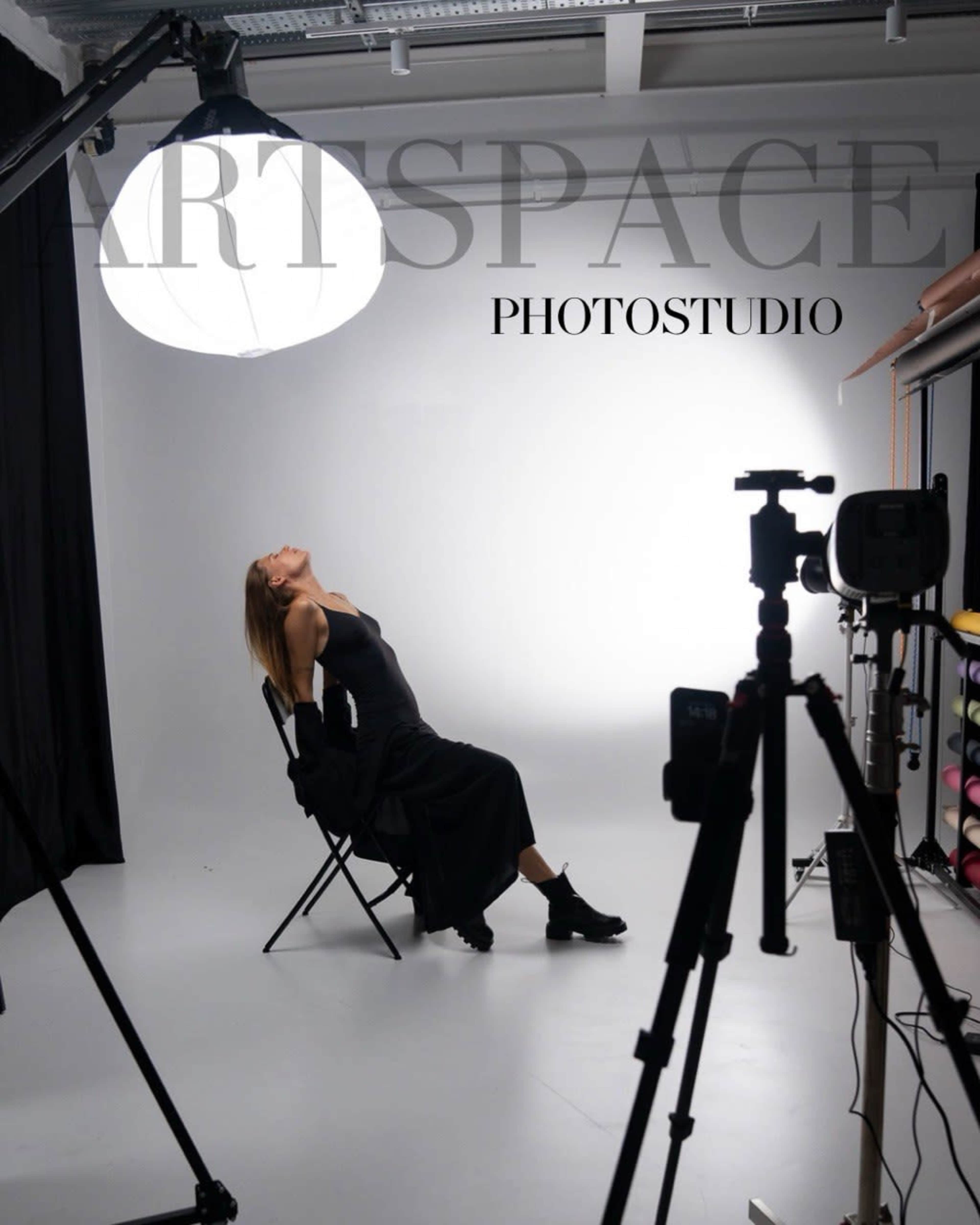 A model poses in a black dress on a chair in a well-lit photography studio, surrounded by equipment.