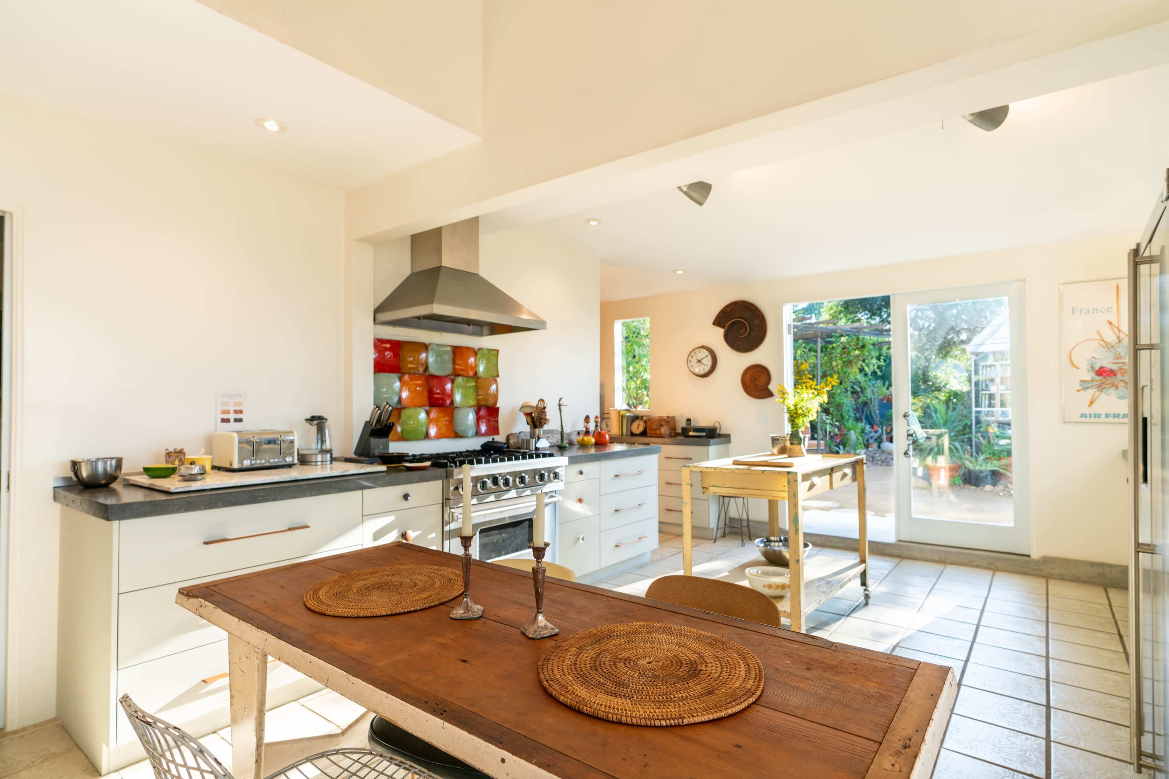A bright kitchen features a central table, a large stove with an oven, and glass doors that open to a garden.