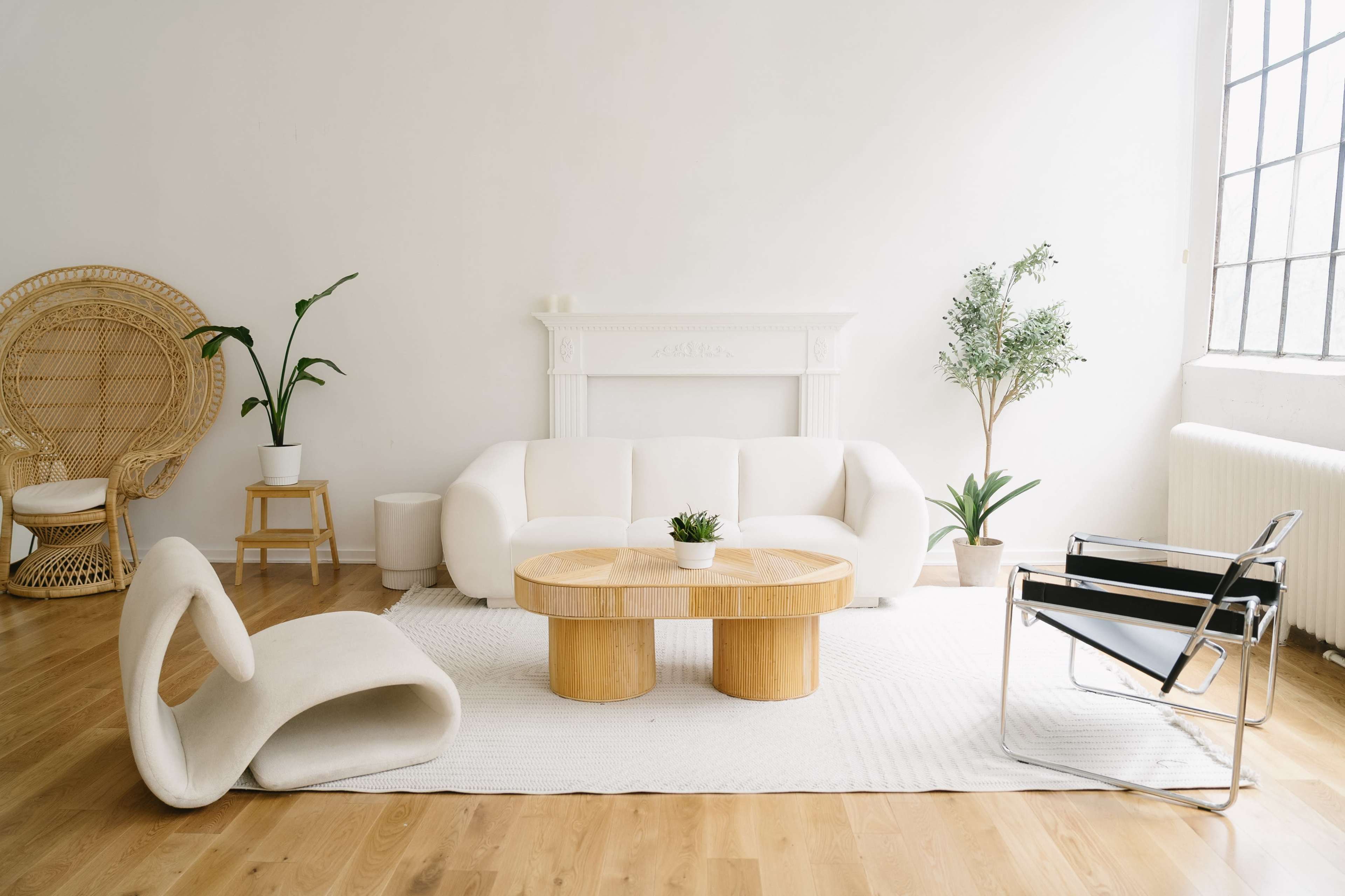 The image shows a modern living room featuring a white sofa, a round wooden coffee table, a pair of indoor plants, and a mix of minimalist furniture.