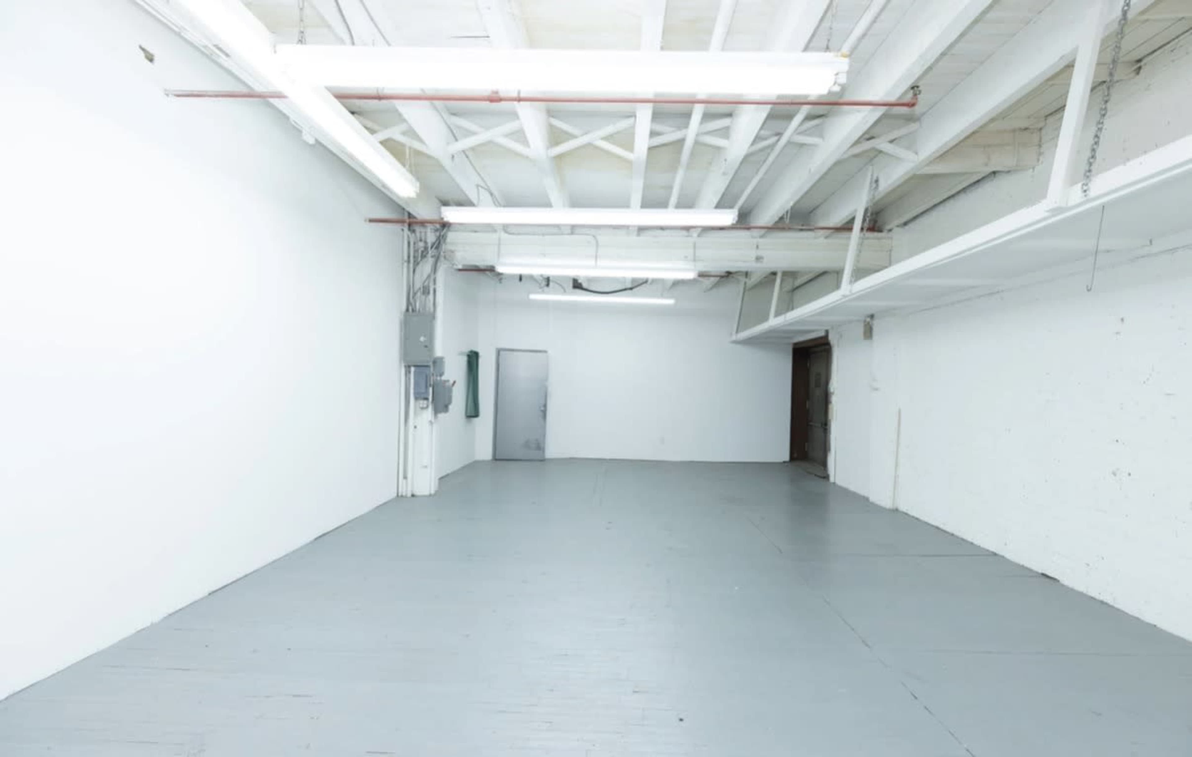 The image shows a spacious, empty room with white walls, a gray floor, and exposed overhead beams.