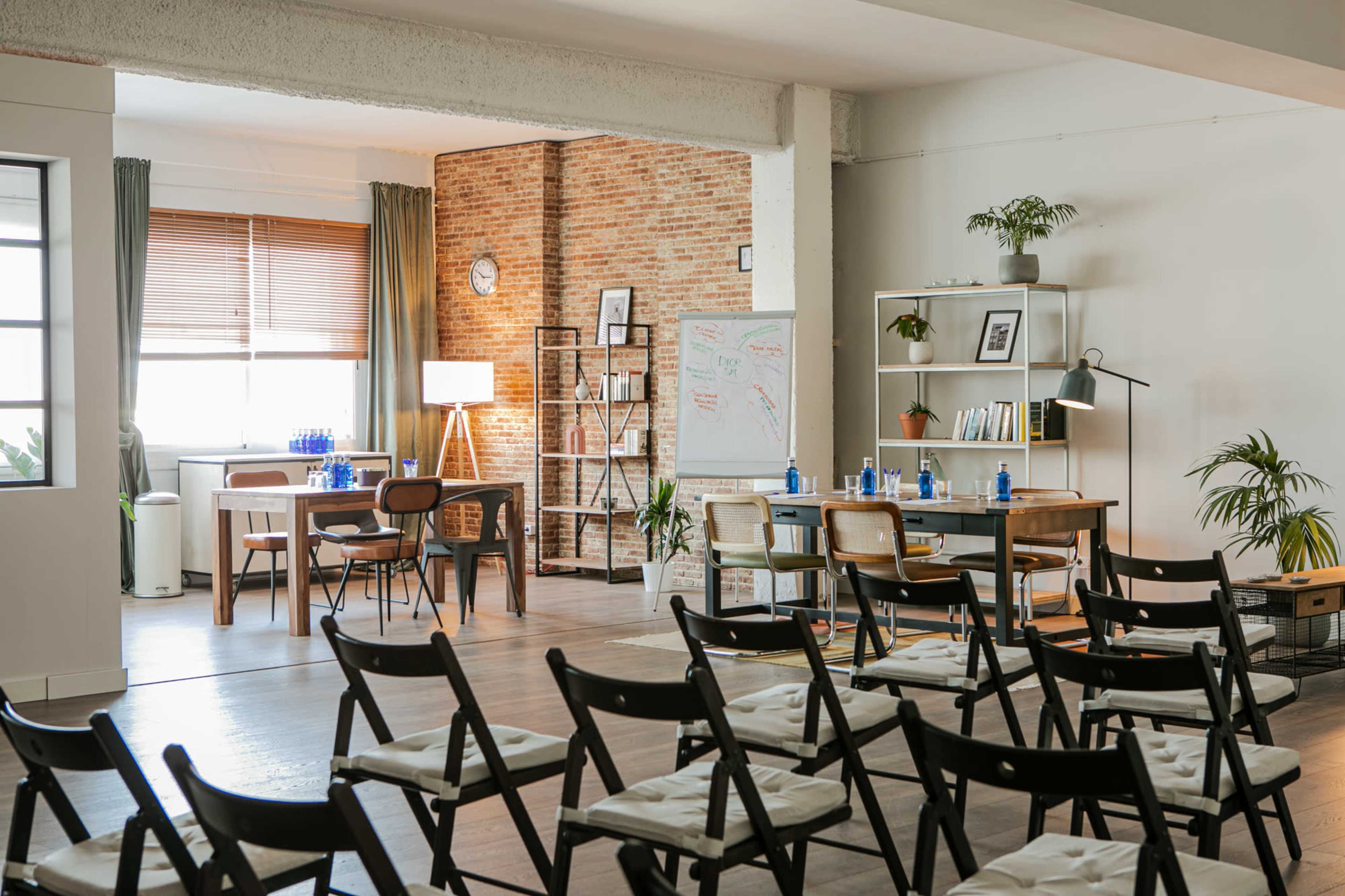 The image shows a modern meeting room with tables, chairs, a whiteboard, and plants, illuminated by natural light.