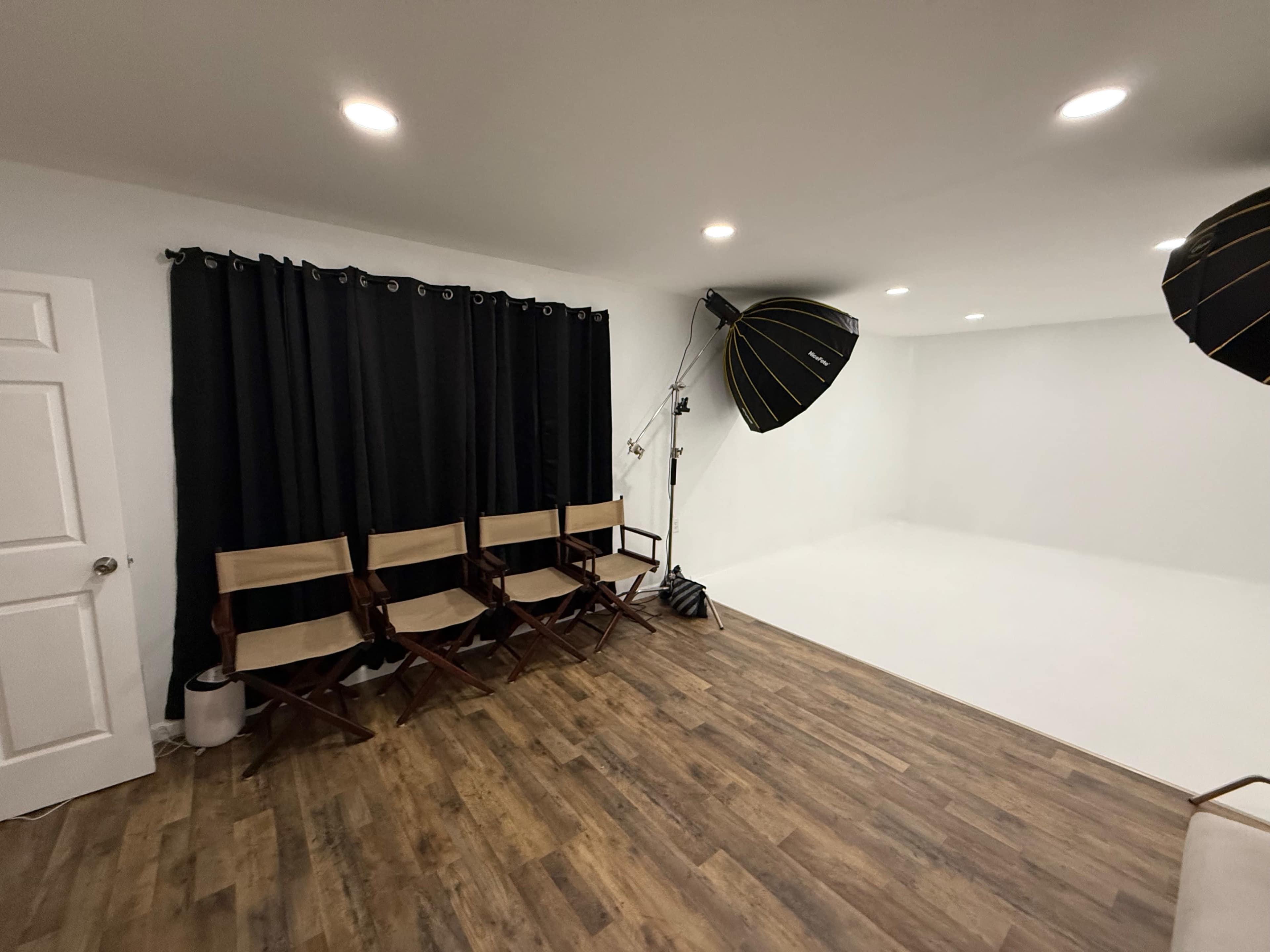 The image shows a photography studio featuring four director's chairs, black curtains, and two large lighting equipment setups, all against a white backdrop.
