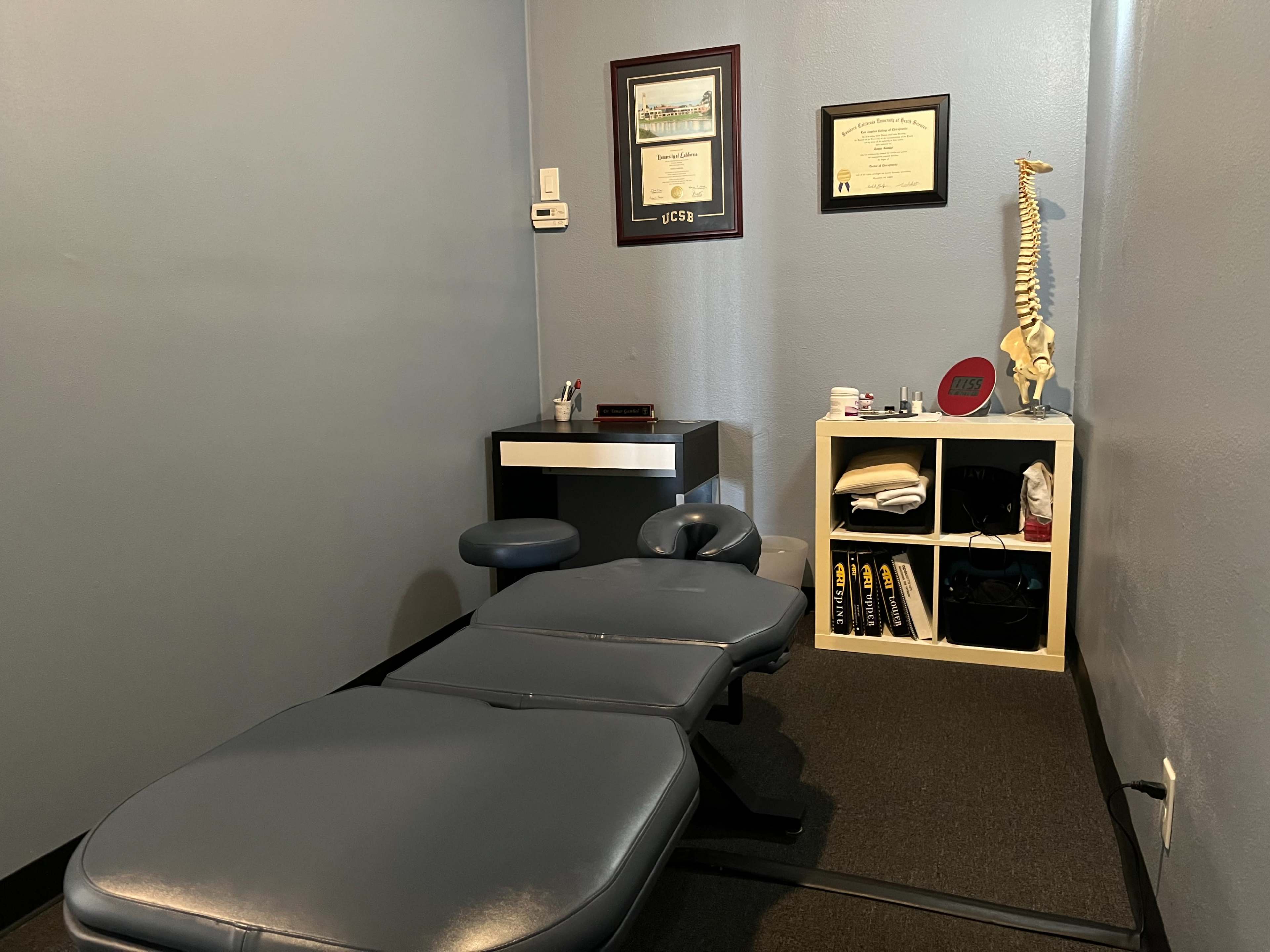 The image shows a medical consultation room featuring a blue examination table, a desk with a chair, shelves with various items, and a framed certificate on the wall.