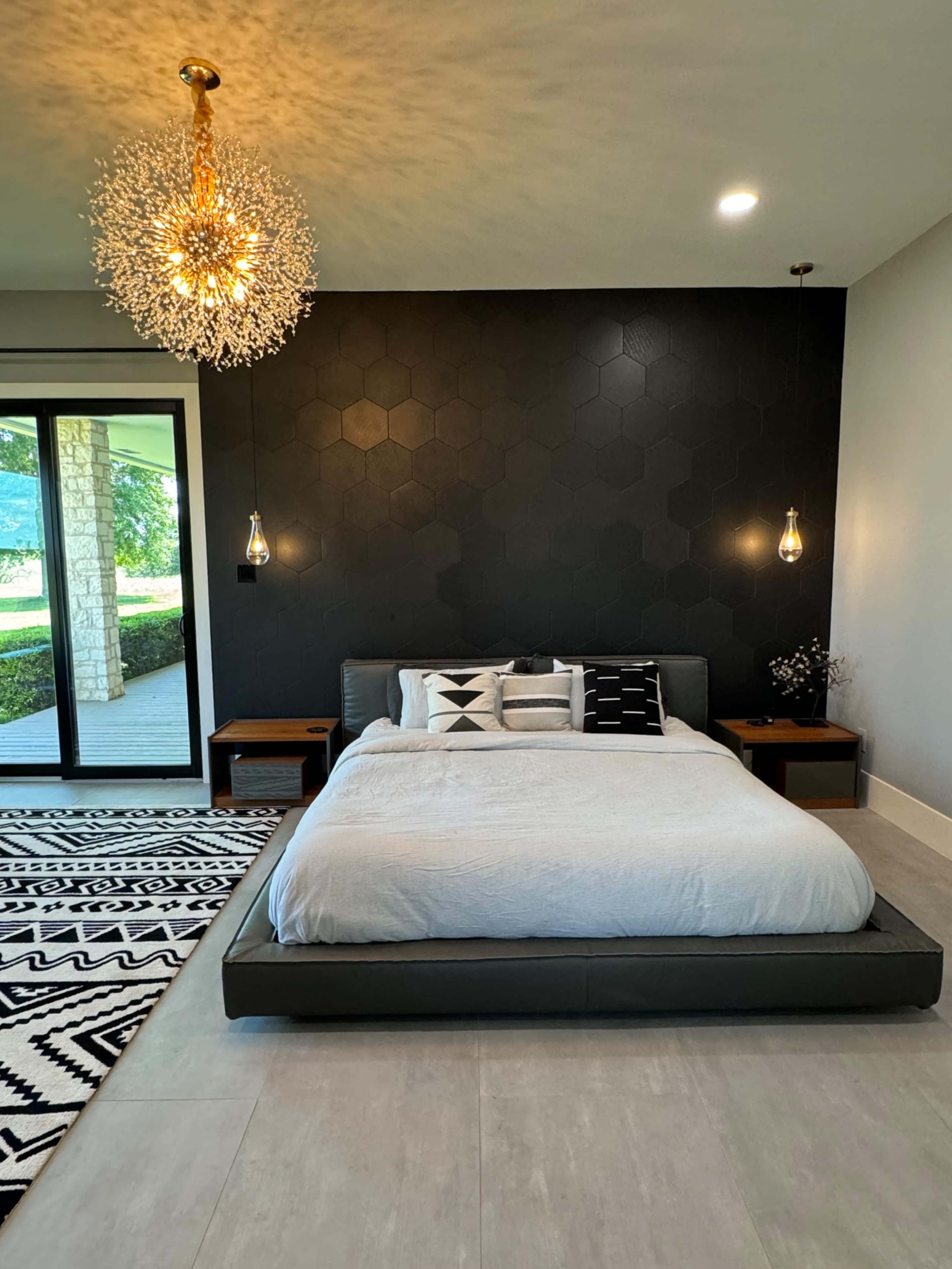 The image shows a modern bedroom featuring a low, minimalist bed with light bedding, geometric-patterned pillows, two pendant lights, and a striking hexagonal accent wall.