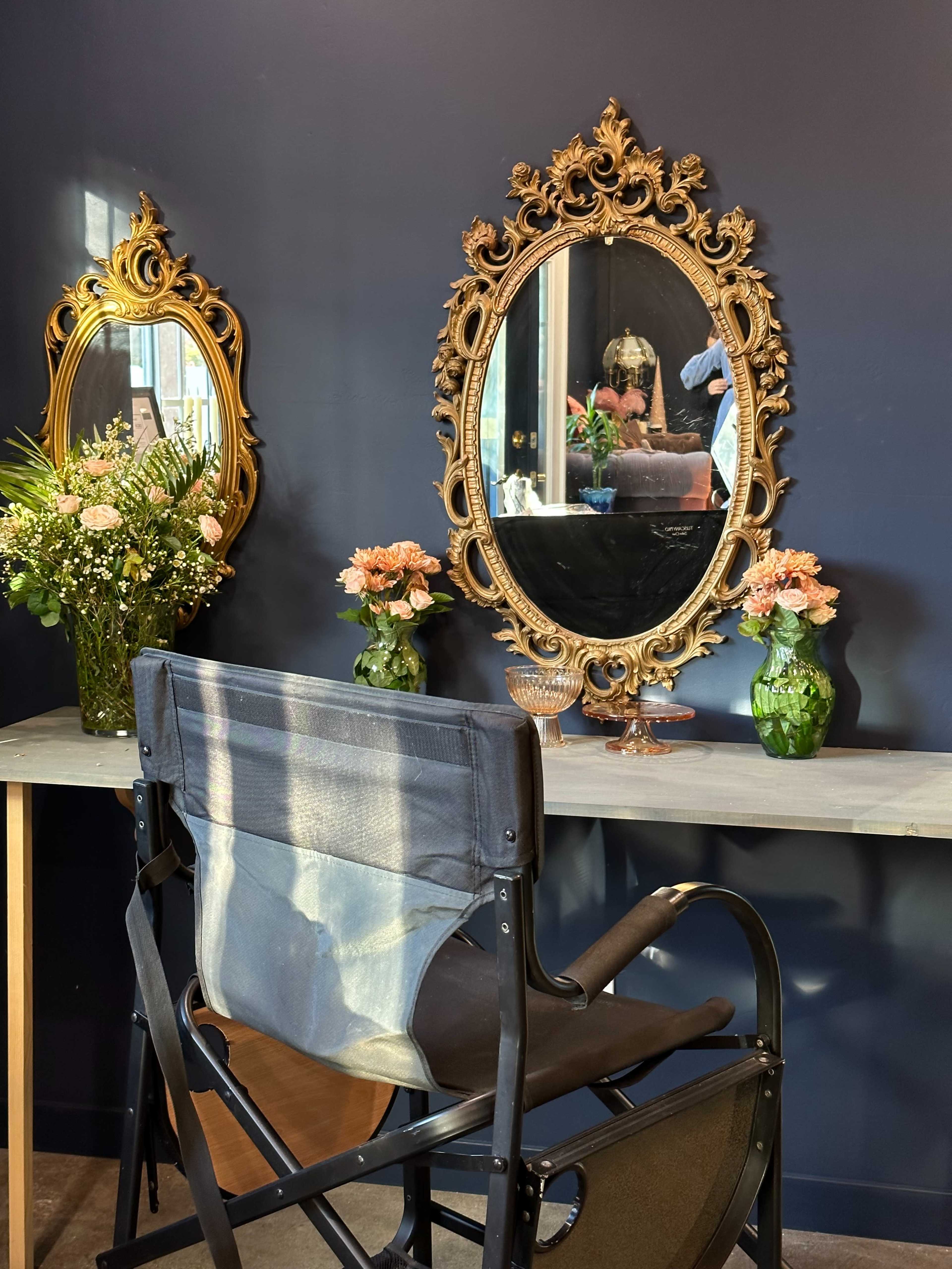 The image features a black wall with two ornate mirrors above a wooden table, flanked by vases of flowers and a director's chair.