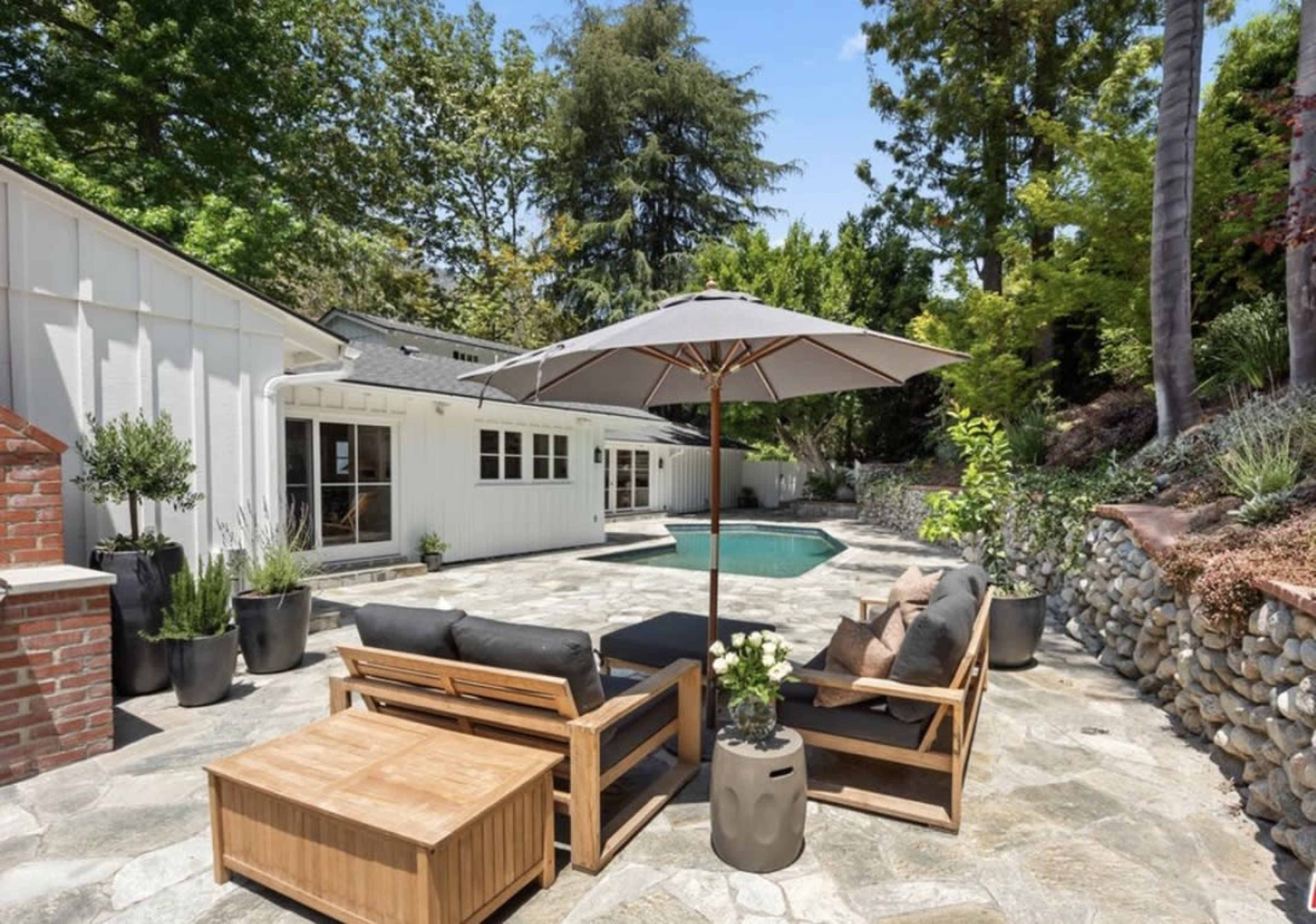 A backyard patio area with a swimming pool, surrounded by stone flooring, wooden furniture, and large planters.