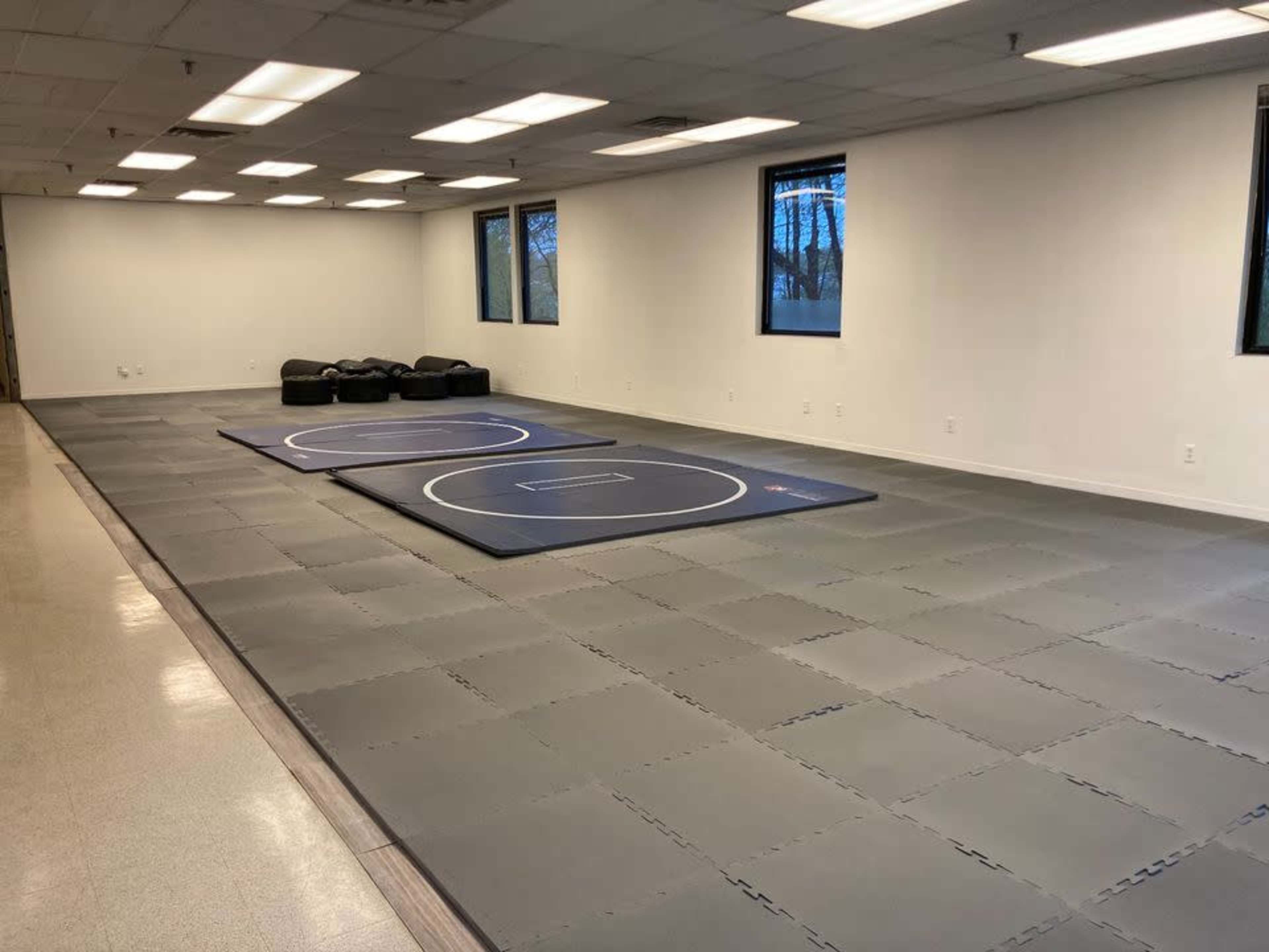 A spacious training room featuring two wrestling mats on a gray tiled floor, with black seating in the background and windows letting in natural light.