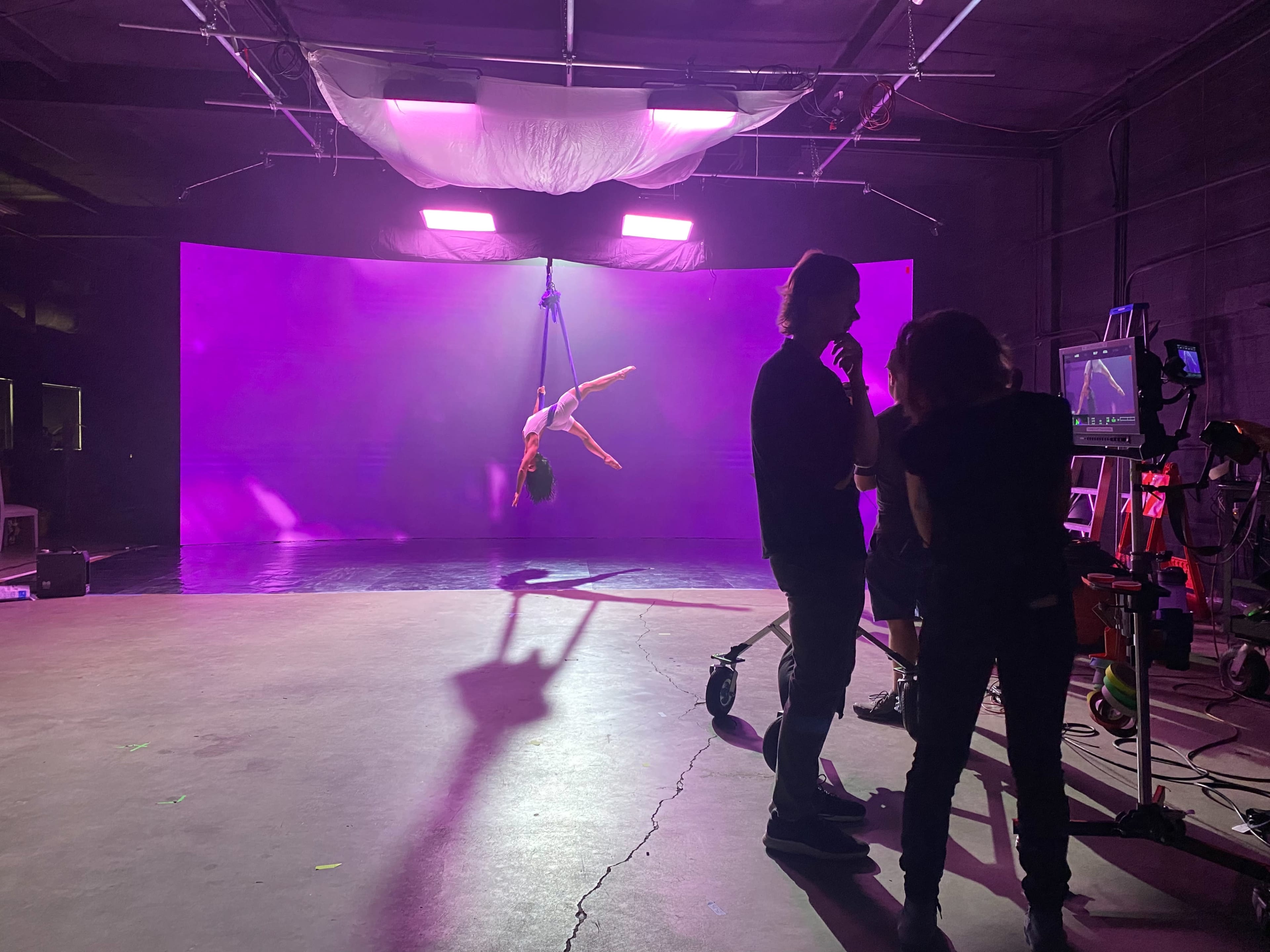 A performer is suspended from the ceiling on a harness against a luminous purple backdrop in a studio setting.