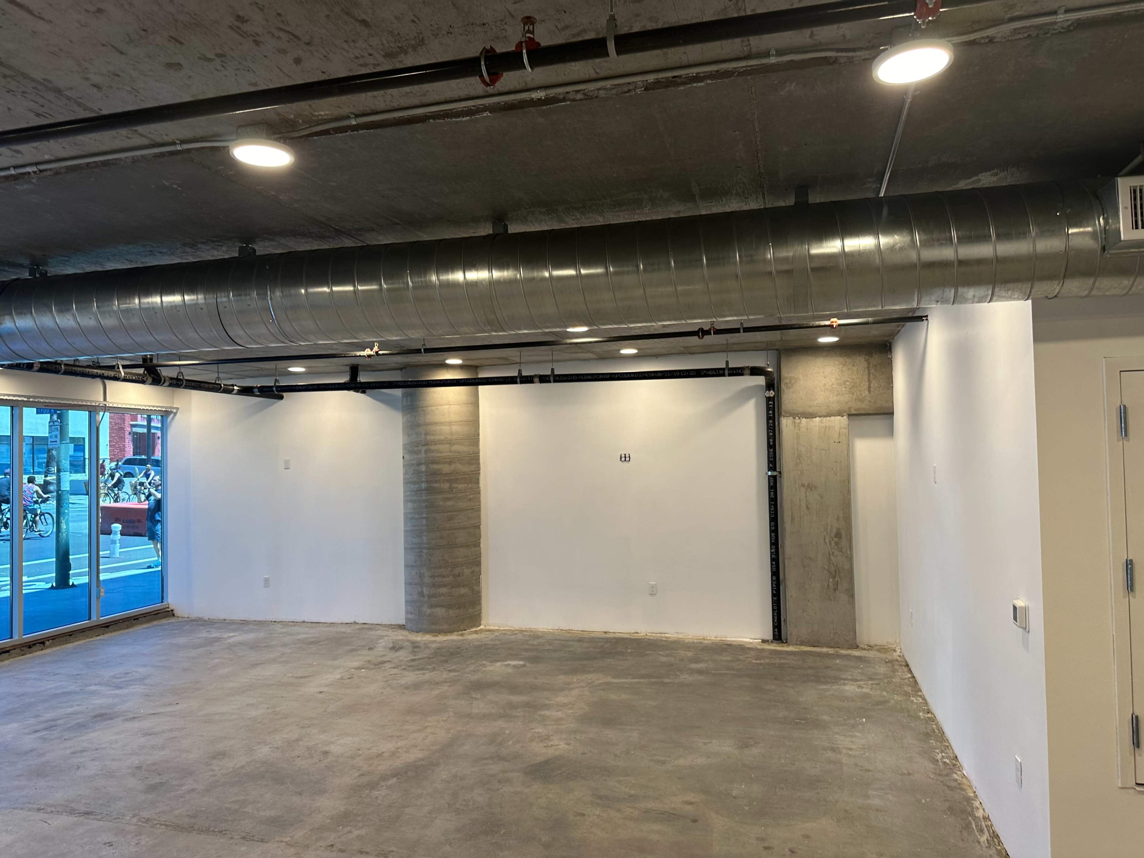 The image shows an unfinished commercial space with exposed concrete walls, a high ceiling, and large windows overlooking the street.