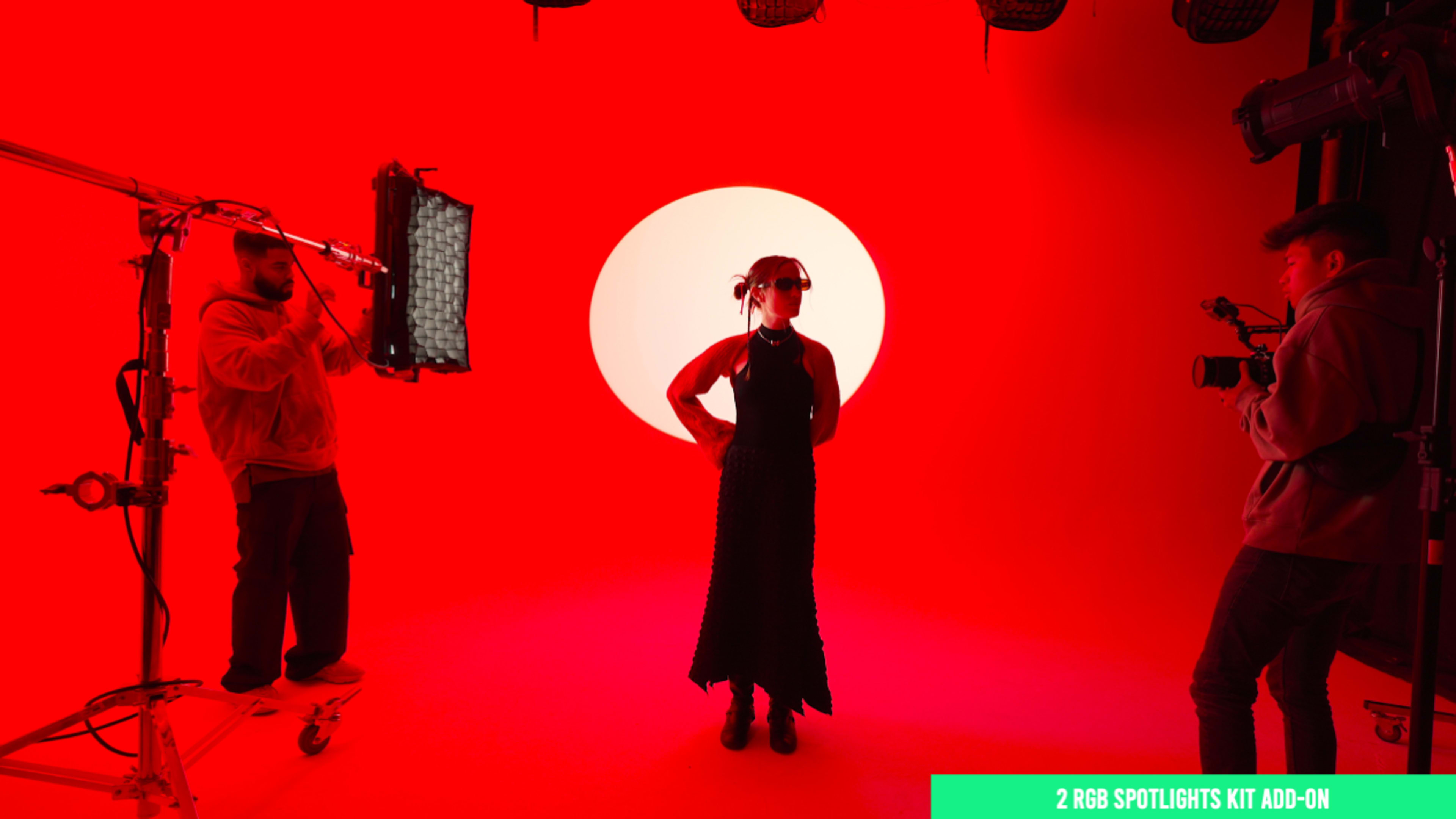 A model stands in front of a large illuminated circle against a red backdrop, while two individuals adjust lighting equipment and film her.
