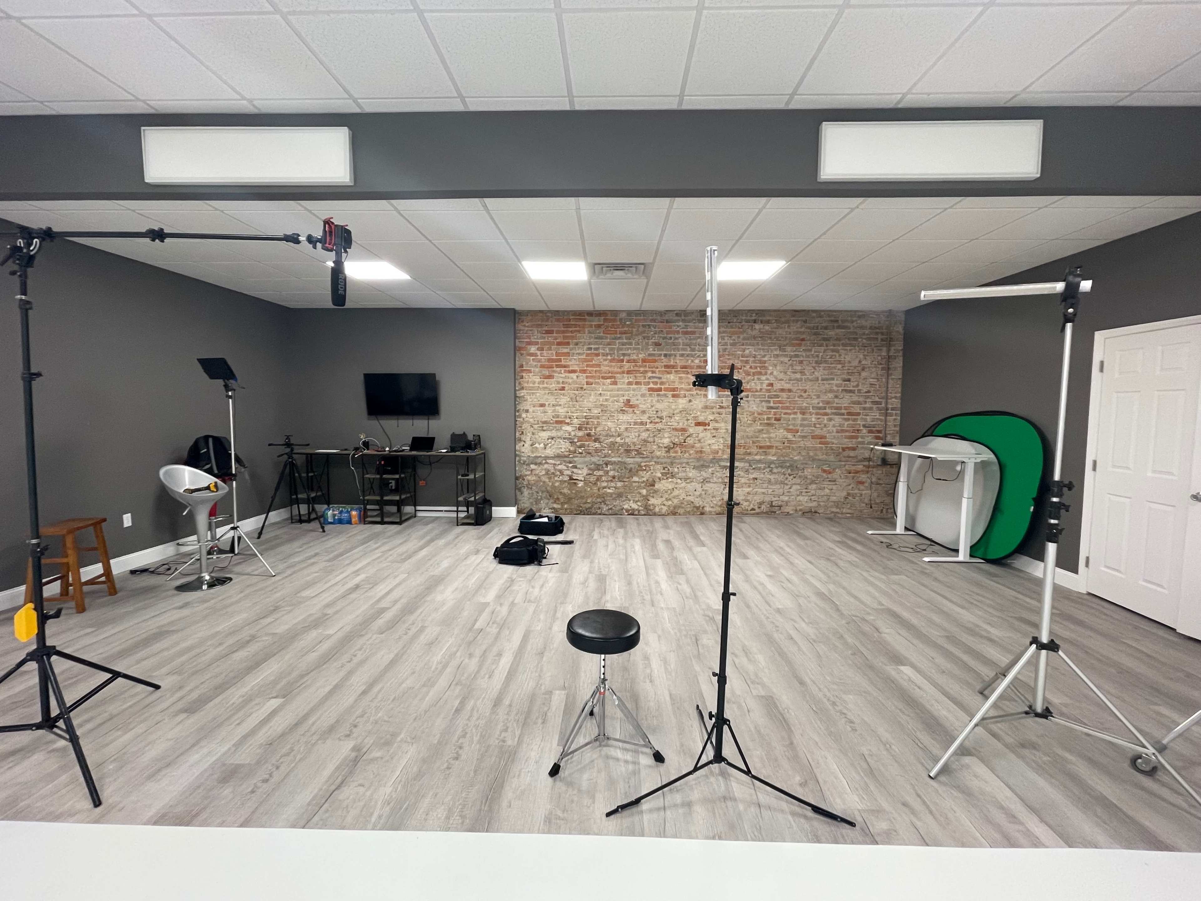 A spacious studio with wooden flooring, featuring several lighting stands, a backdrop area with a green screen, and equipment storage along a brick wall.