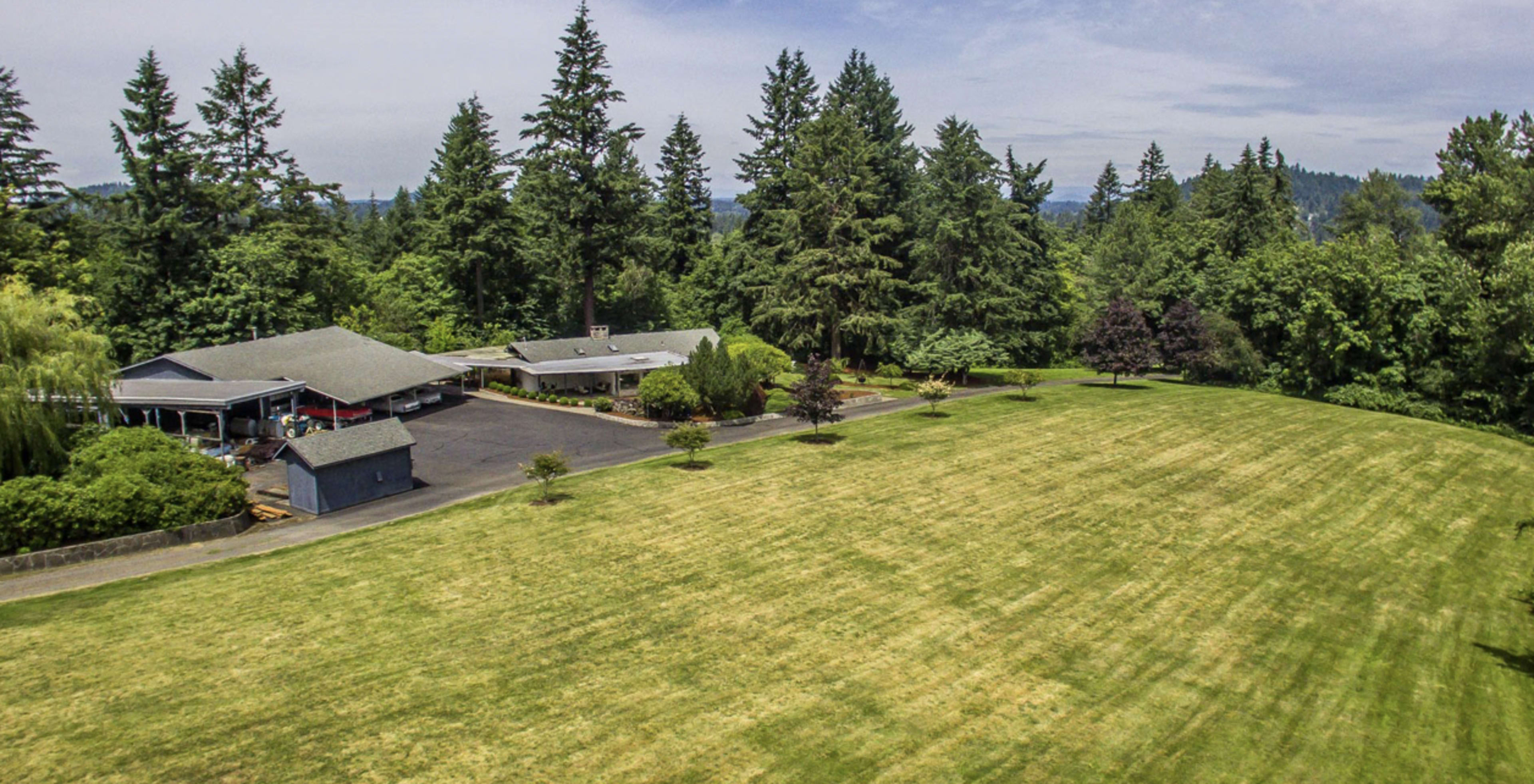 A sprawling house is situated on a grassy lawn surrounded by dense trees in a rural setting.