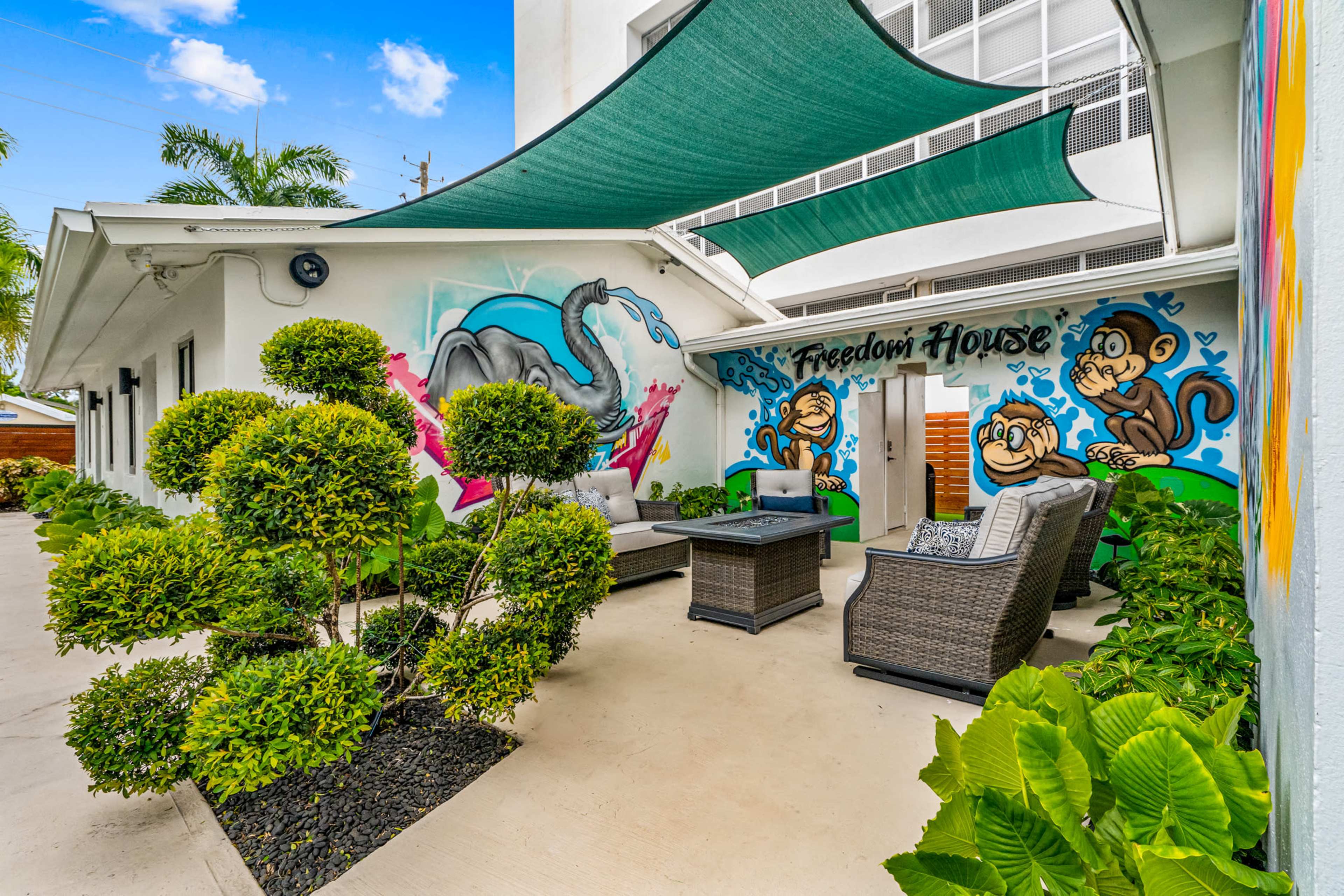 The image shows a courtyard with a mural featuring monkeys on the wall, surrounded by green plants and outdoor seating under a shaded area.