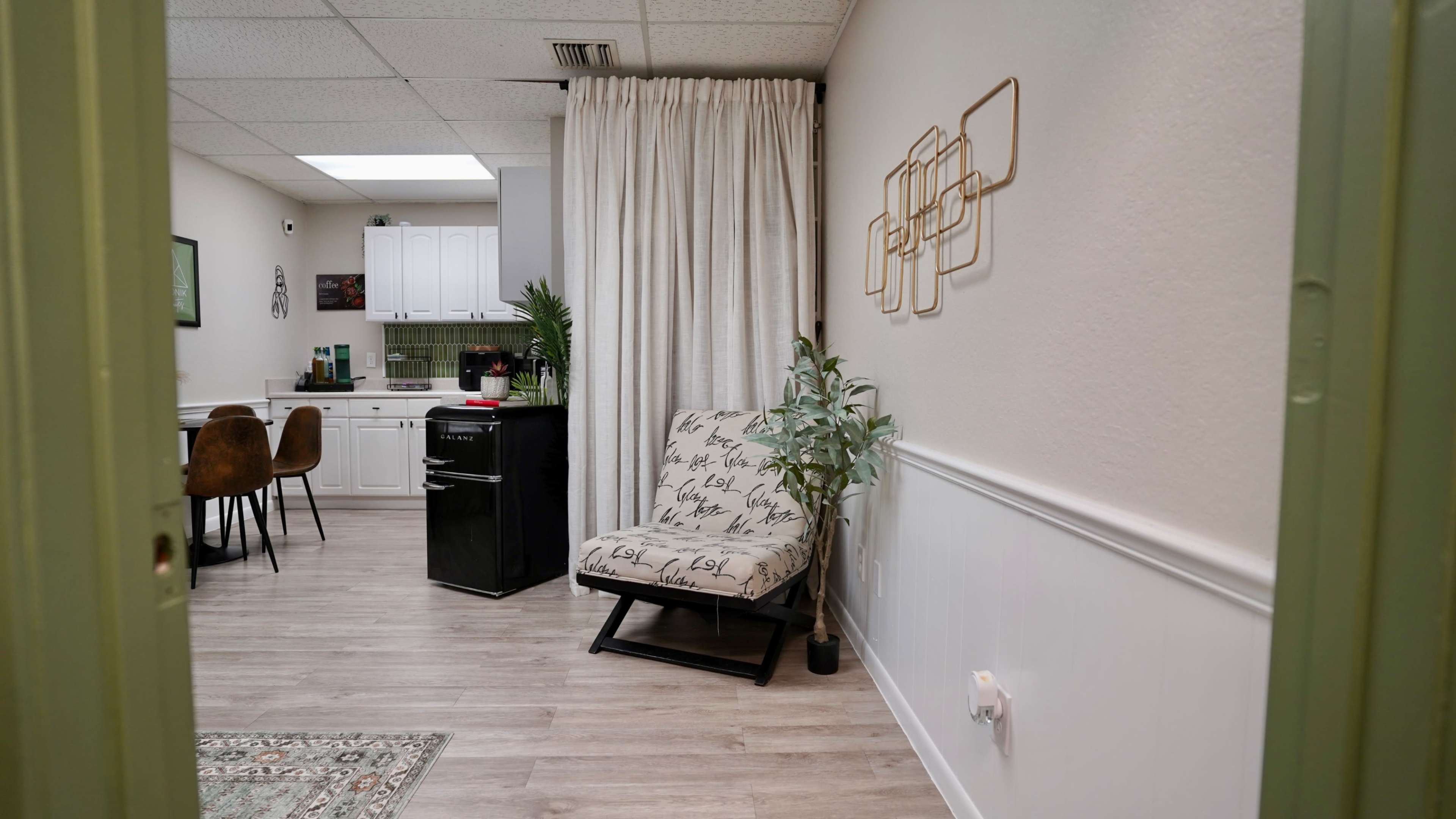 The image shows a small interior space featuring a kitchenette, a folding chair with a patterned cushion, a plant, and decorative wall art.