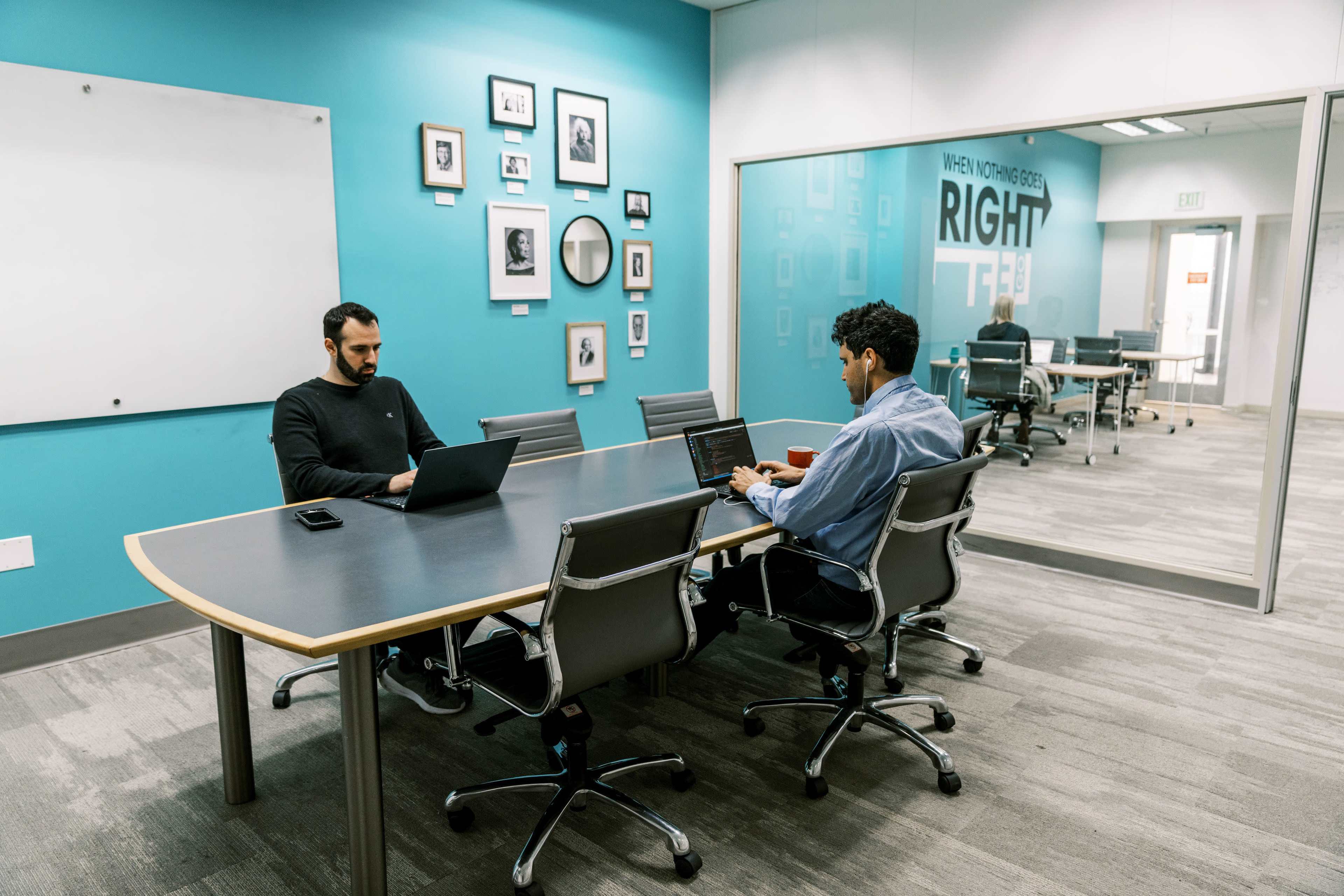 Two men are seated at a conference table, each working on laptops in a brightly colored meeting room.