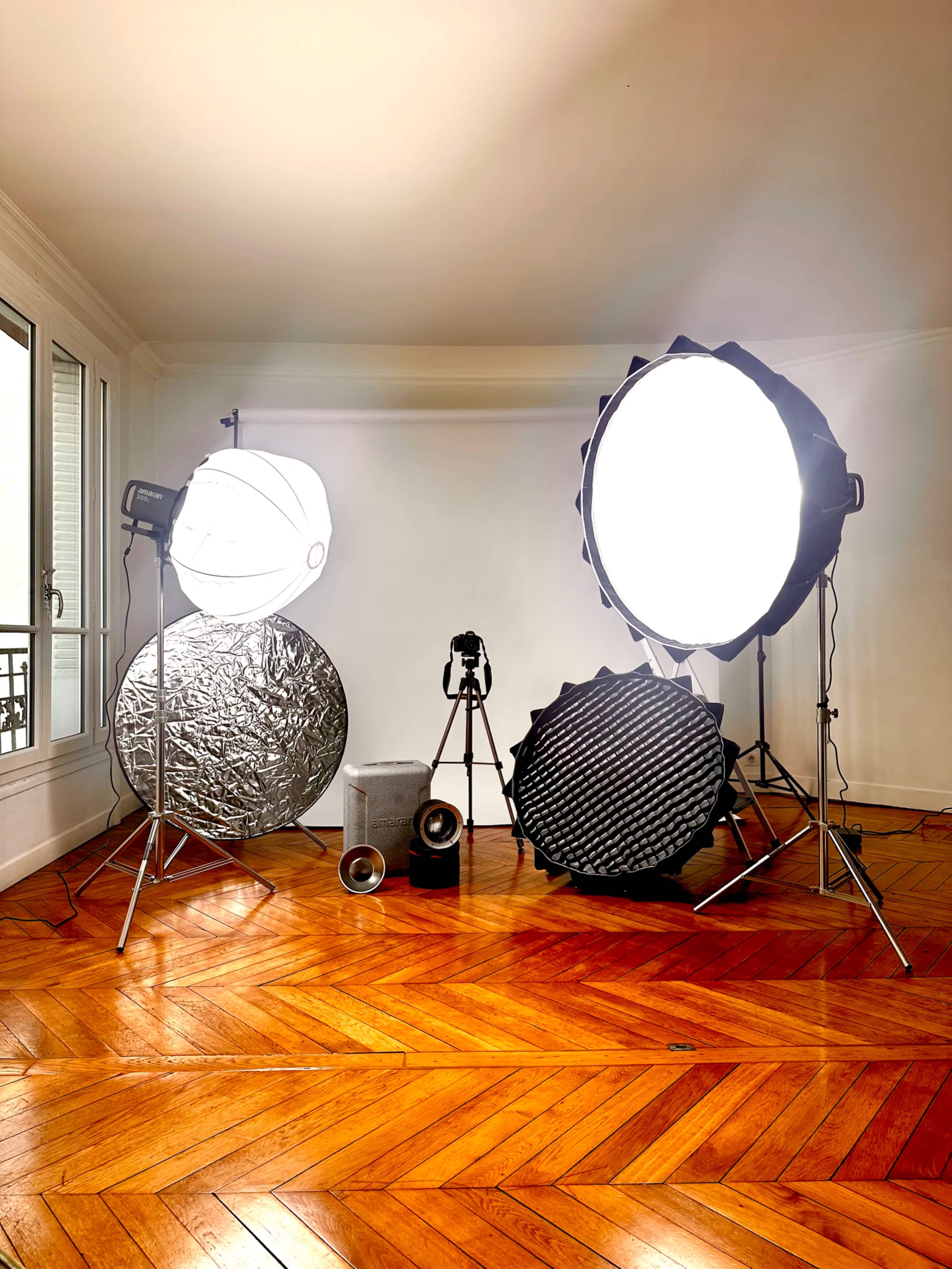 The image shows a photography setup in a room featuring several light sources, reflectors, and a camera on a tripod arranged on a wooden floor.