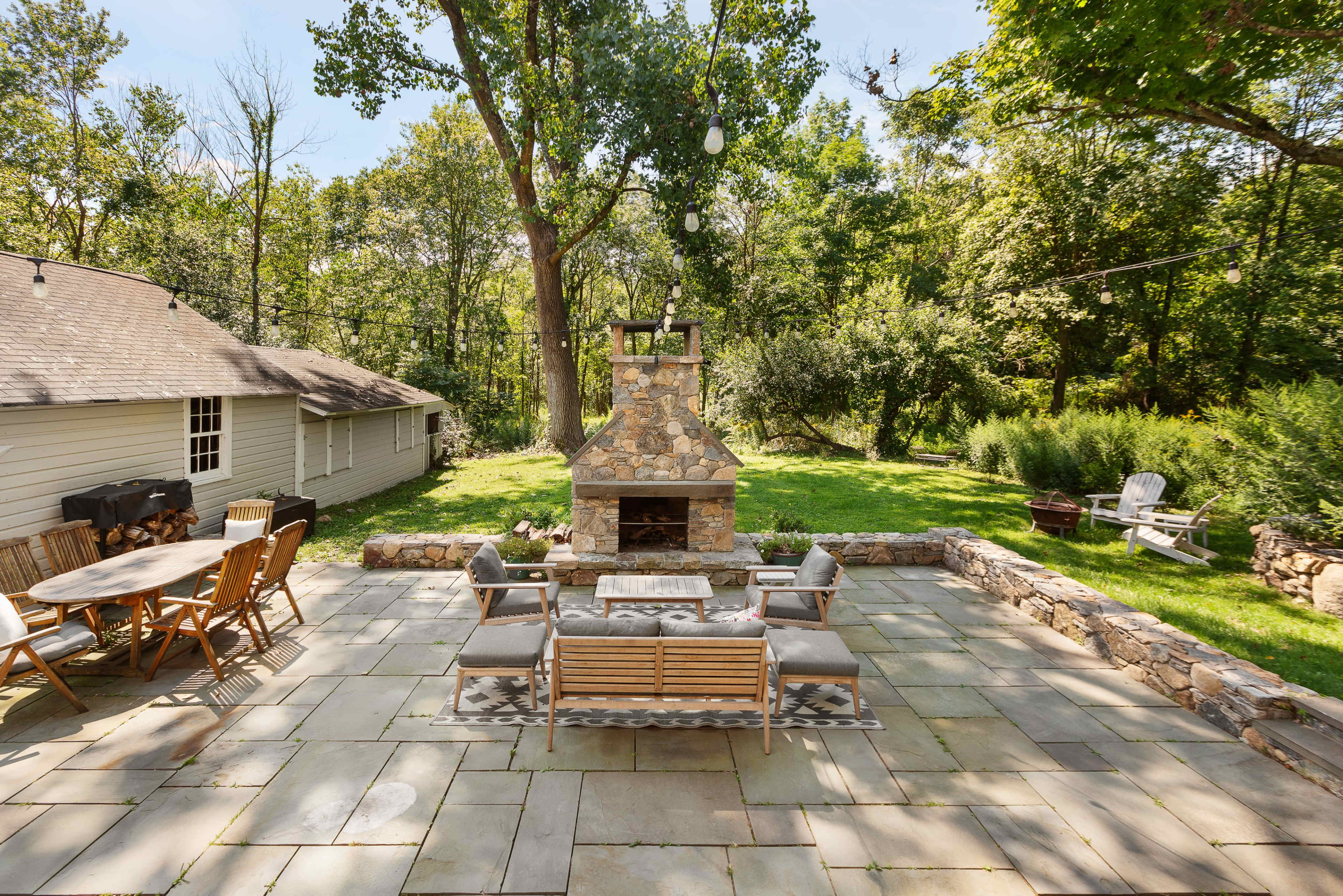 A stone-paved patio with a central fireplace, surrounded by wooden furniture and lush greenery.