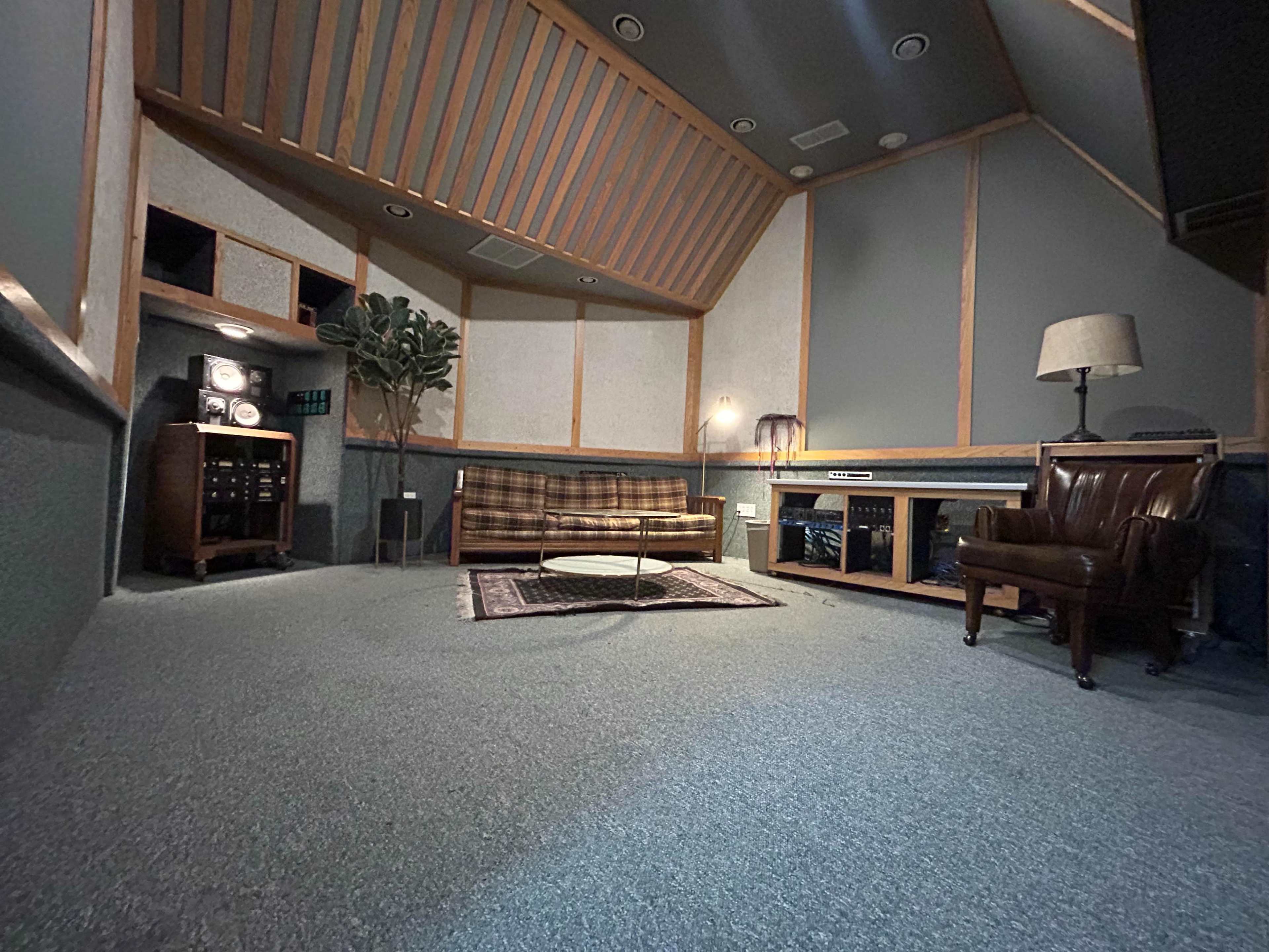 The image shows a recording studio lounge area with a patterned couch, a wooden table, and a leather armchair, surrounded by soundproof walls and a high, sloped ceiling.