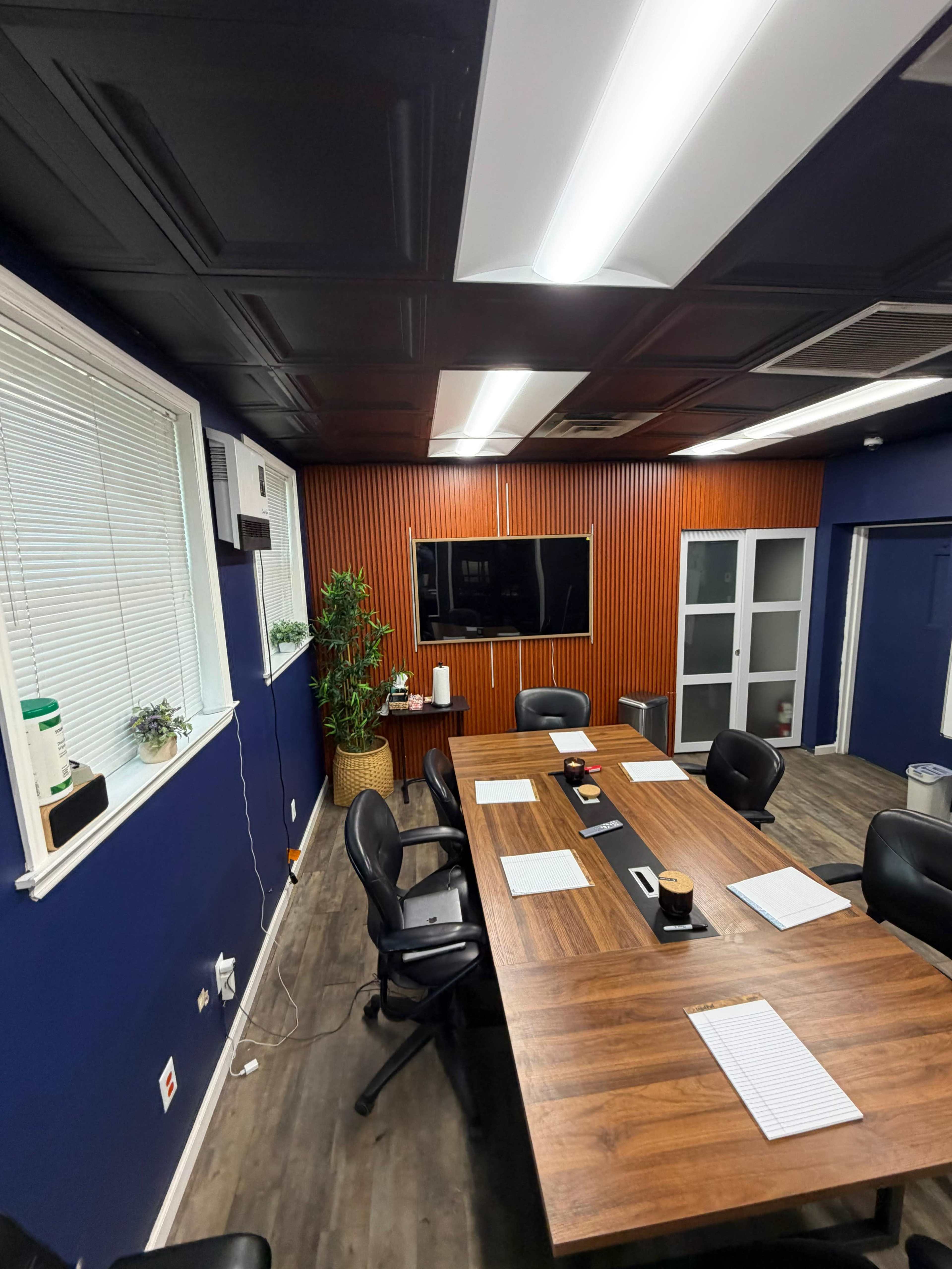 A conference room features a wooden table surrounded by black chairs, with a television mounted on the wall and large windows allowing natural light.
