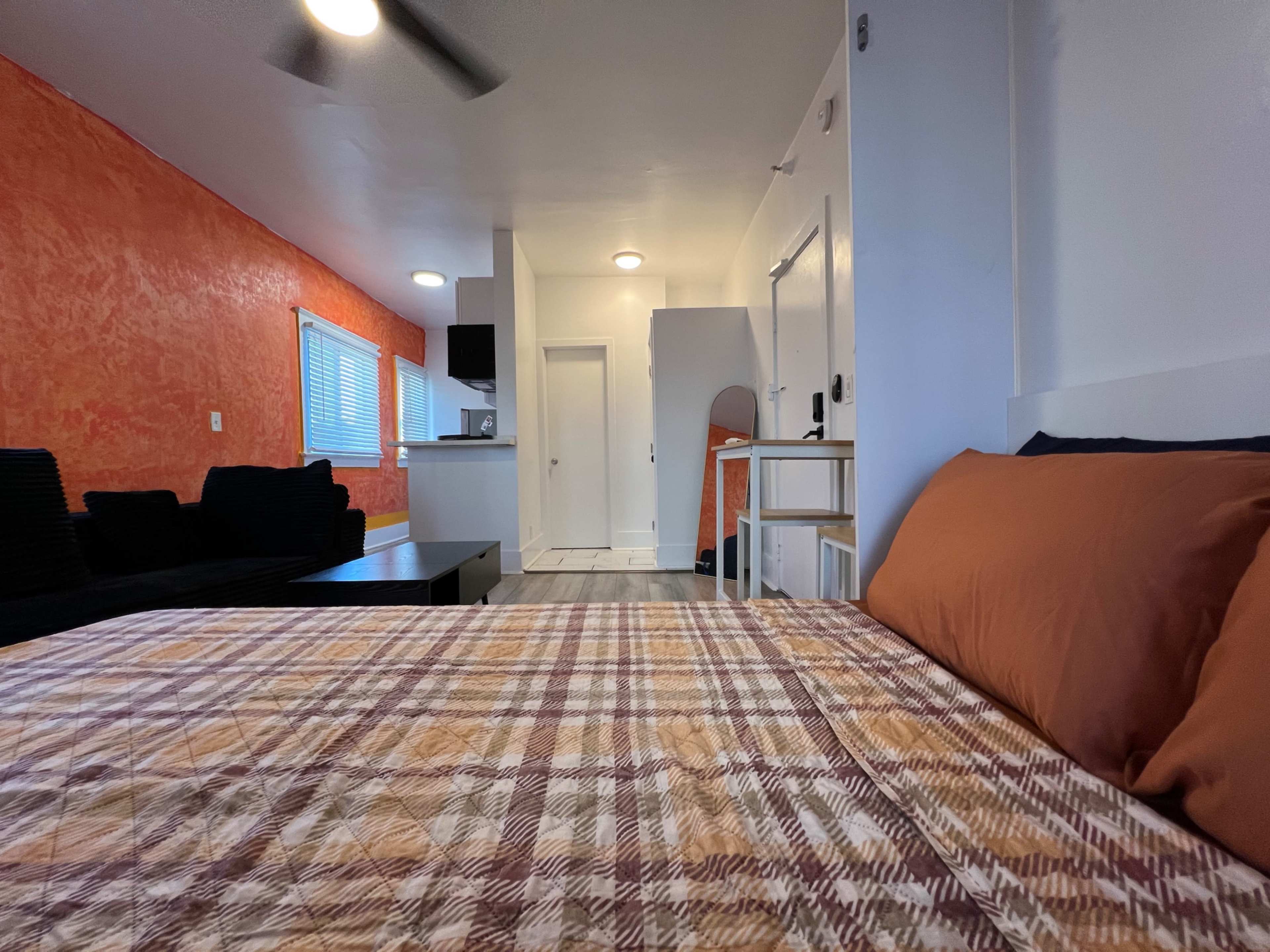 The image shows a small interior space featuring a bedroom with a plaid bedspread, a kitchenette area, and a living area with a black couch against an orange wall.