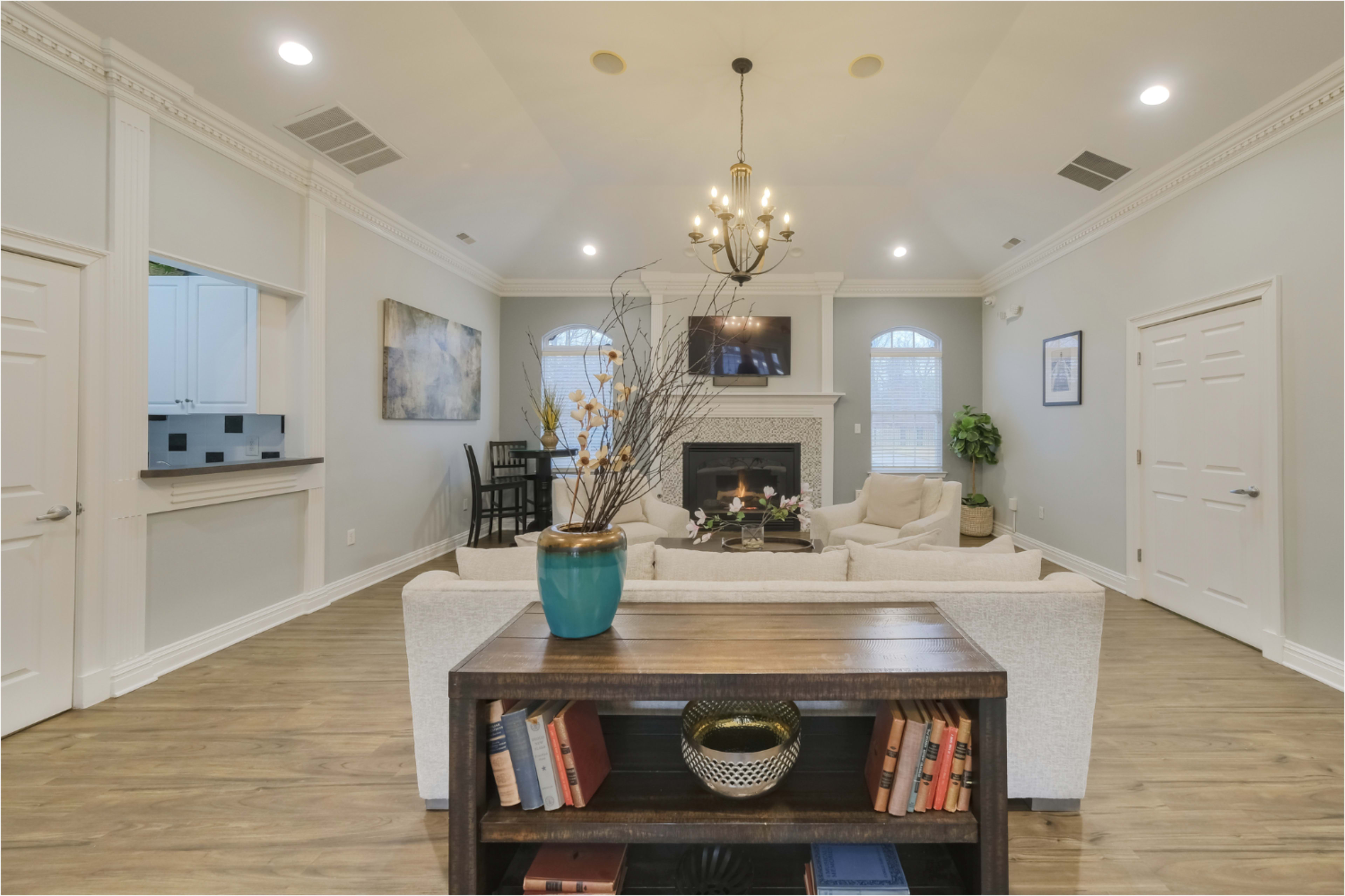 The image shows a bright living room with a fireplace, large windows, and a cozy seating arrangement centered around a coffee table, accented by decorative elements on a wooden shelf.