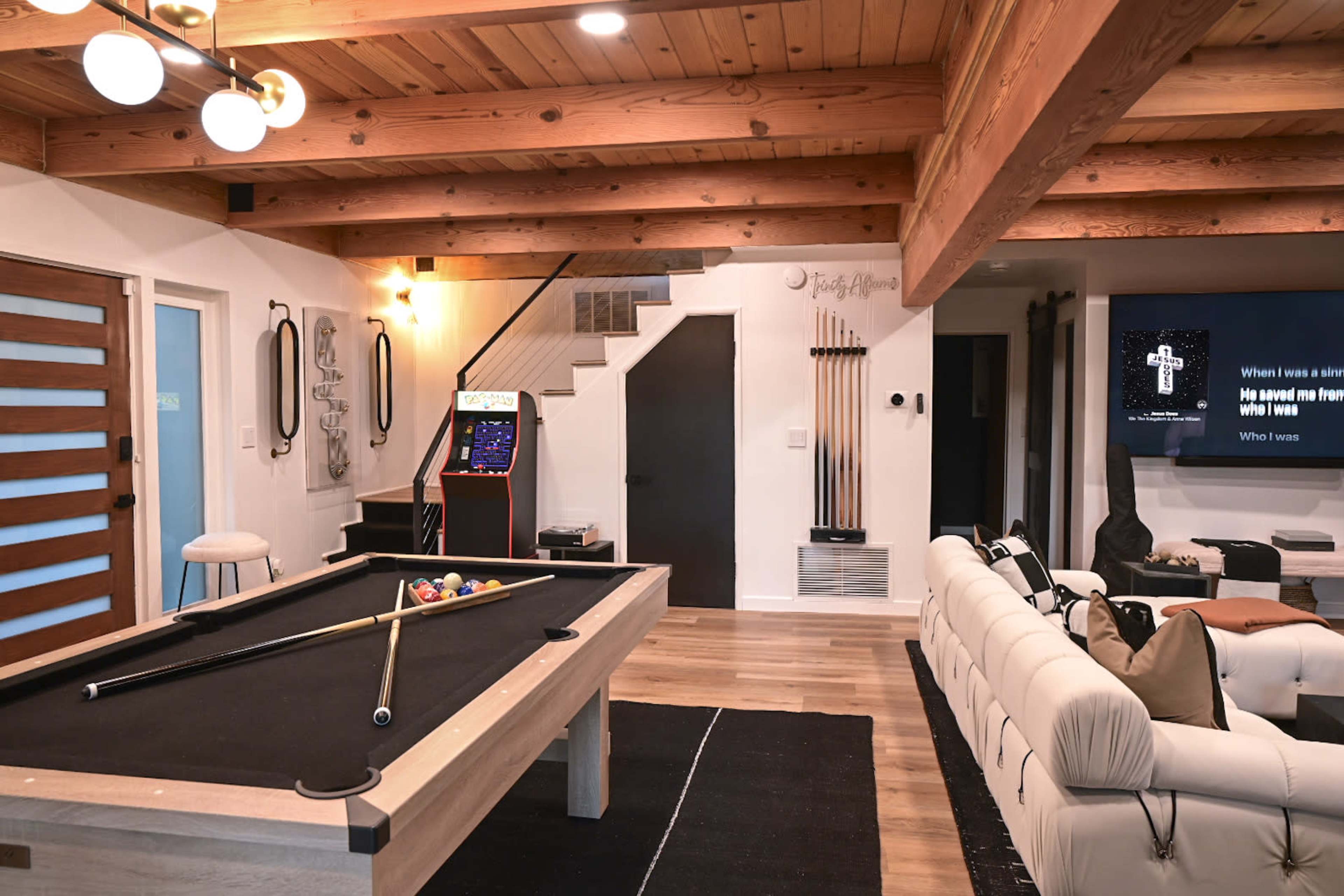 A modern game room featuring a pool table, arcade game machine, and a wall-mounted television, all set within a space with wooden beams and a neutral color palette.