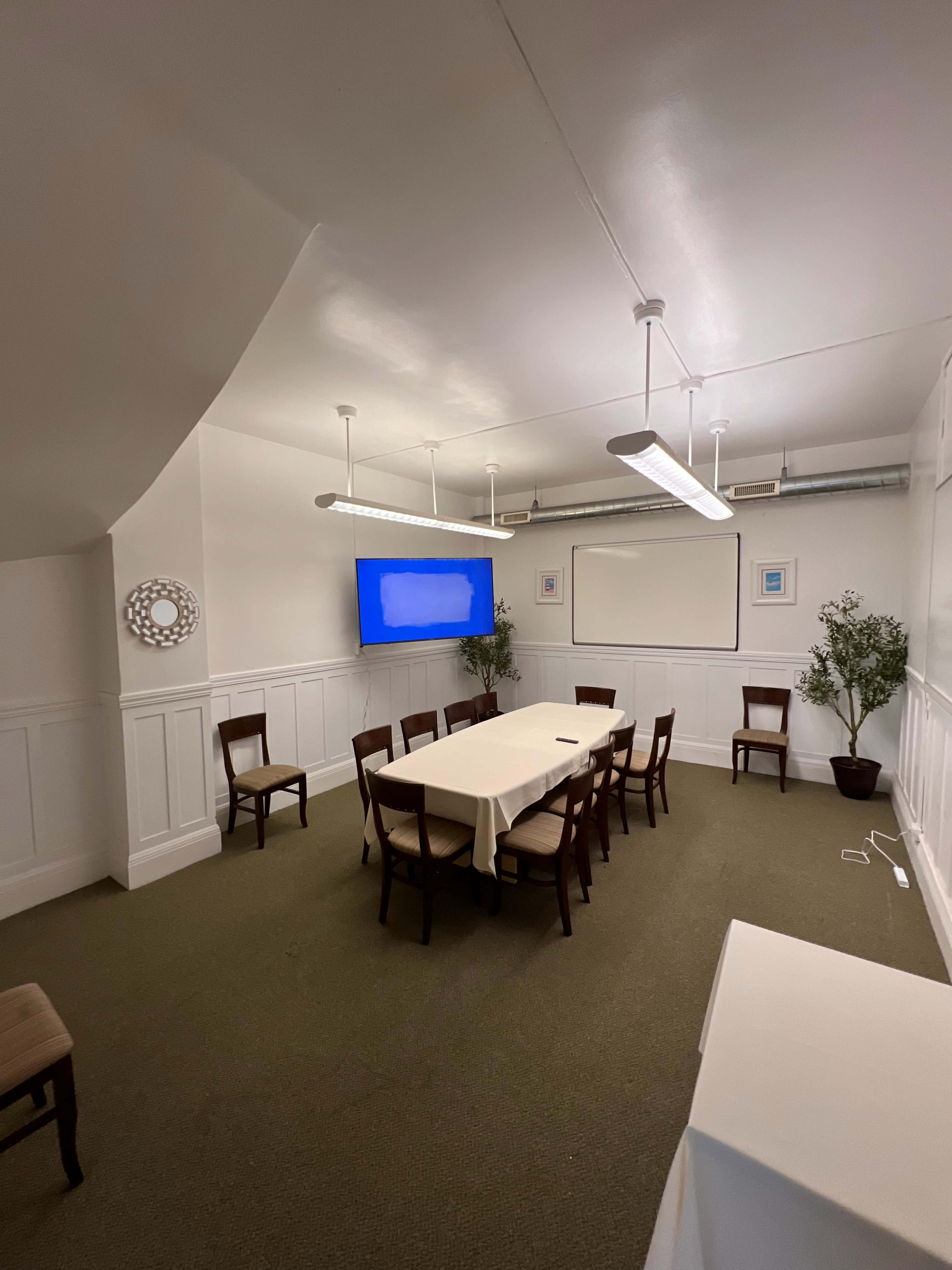 The image shows a meeting room with a rectangular table set for twelve chairs, a large screen on one wall, and a couple of potted plants in the corners.