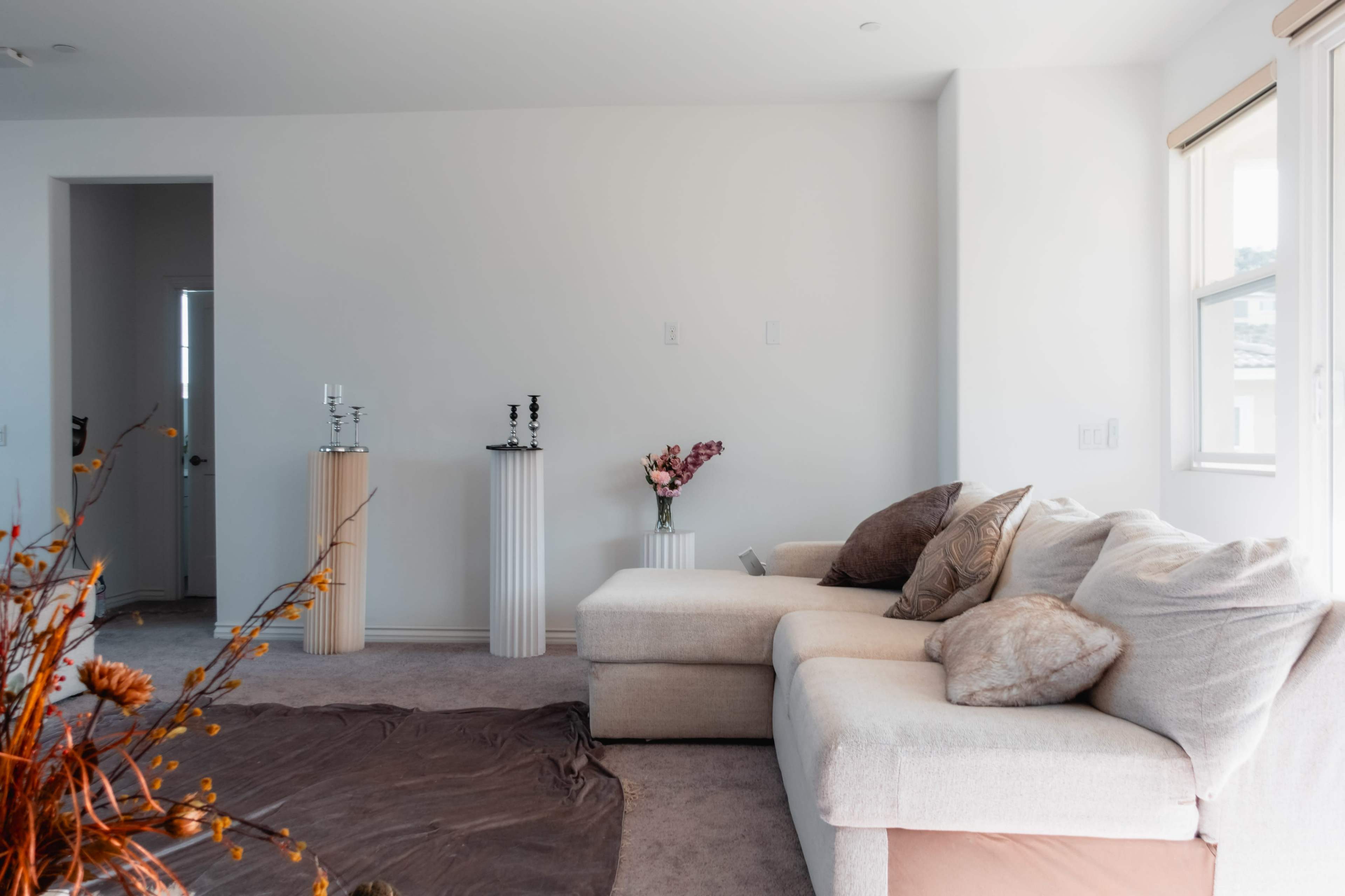 A light-filled living room features a beige sectional sofa, a thick brown rug, and two decorative columns with floral arrangements.
