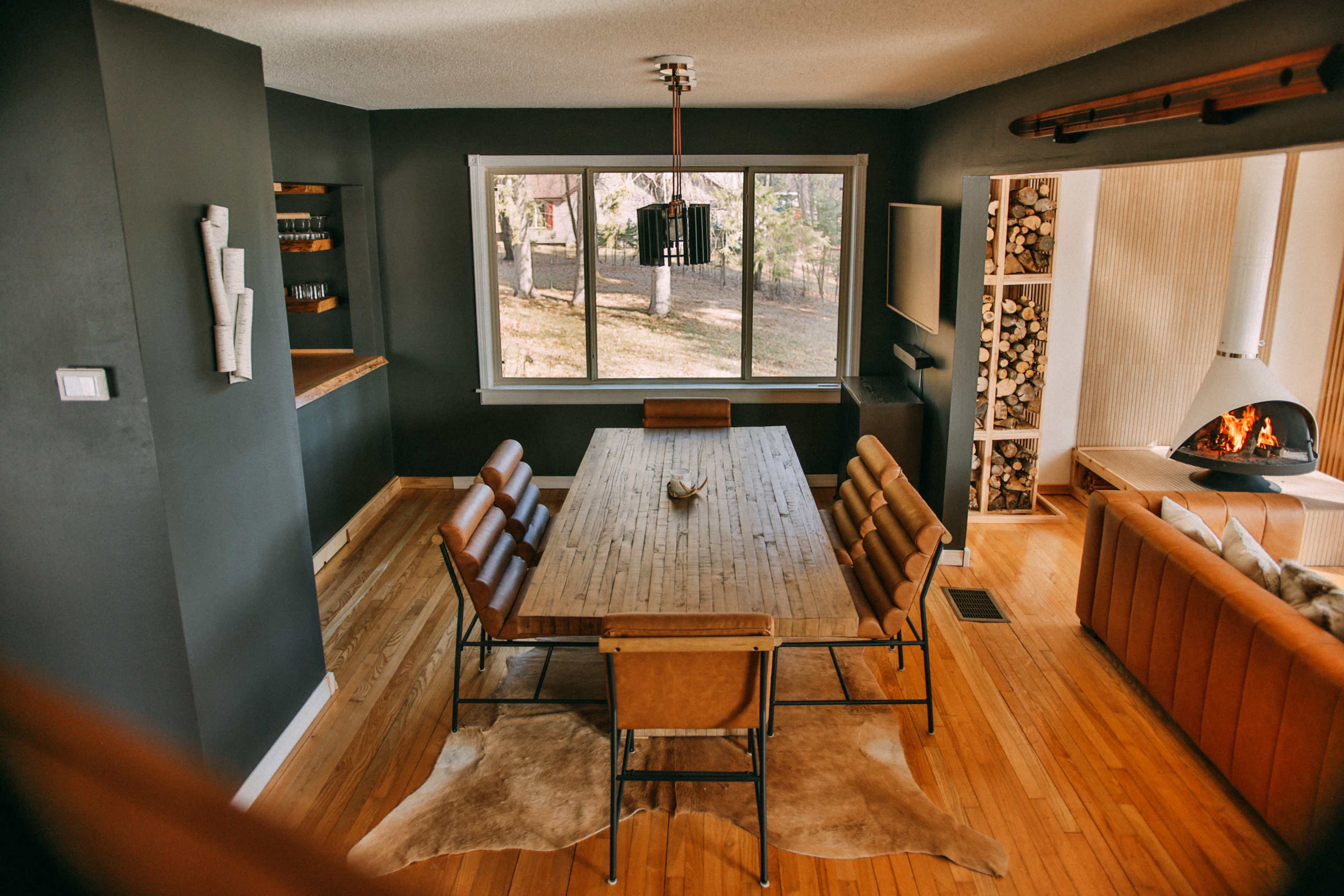 The image shows a modern dining area featuring a wooden table surrounded by leather chairs, large windows providing natural light, and a cozy fireplace with stacked logs nearby.