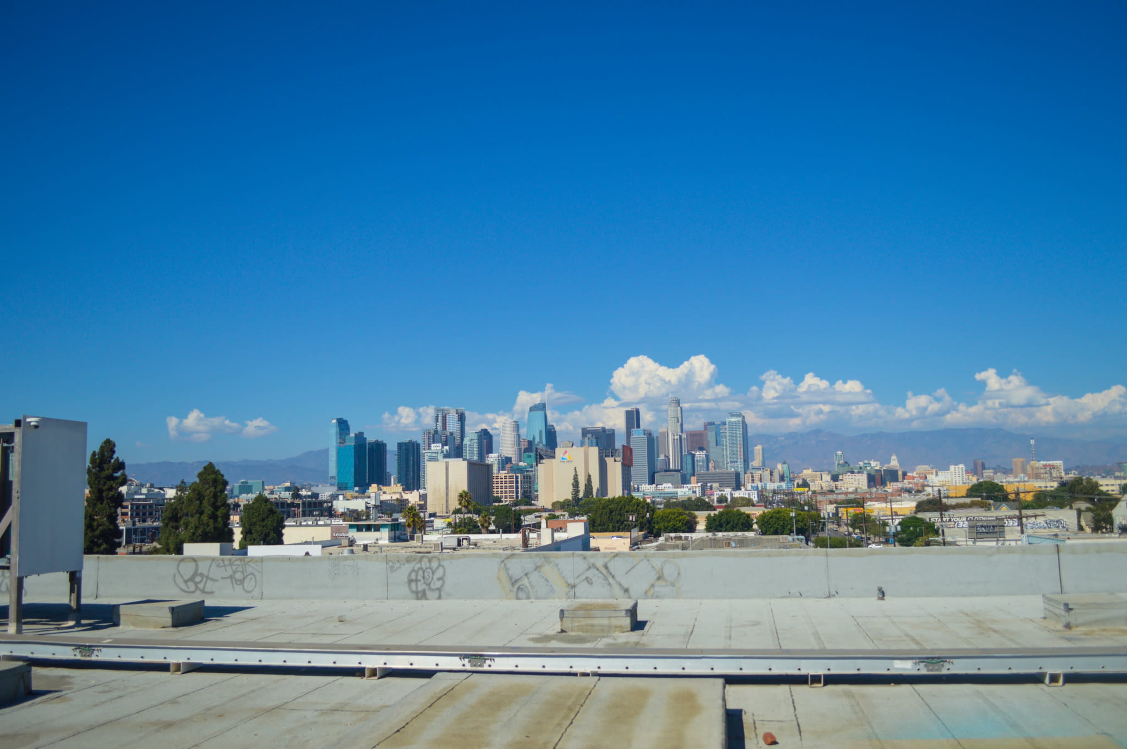 A clear view from a rooftop overlooks the skyline of downtown Los Angeles against a blue sky with scattered clouds.