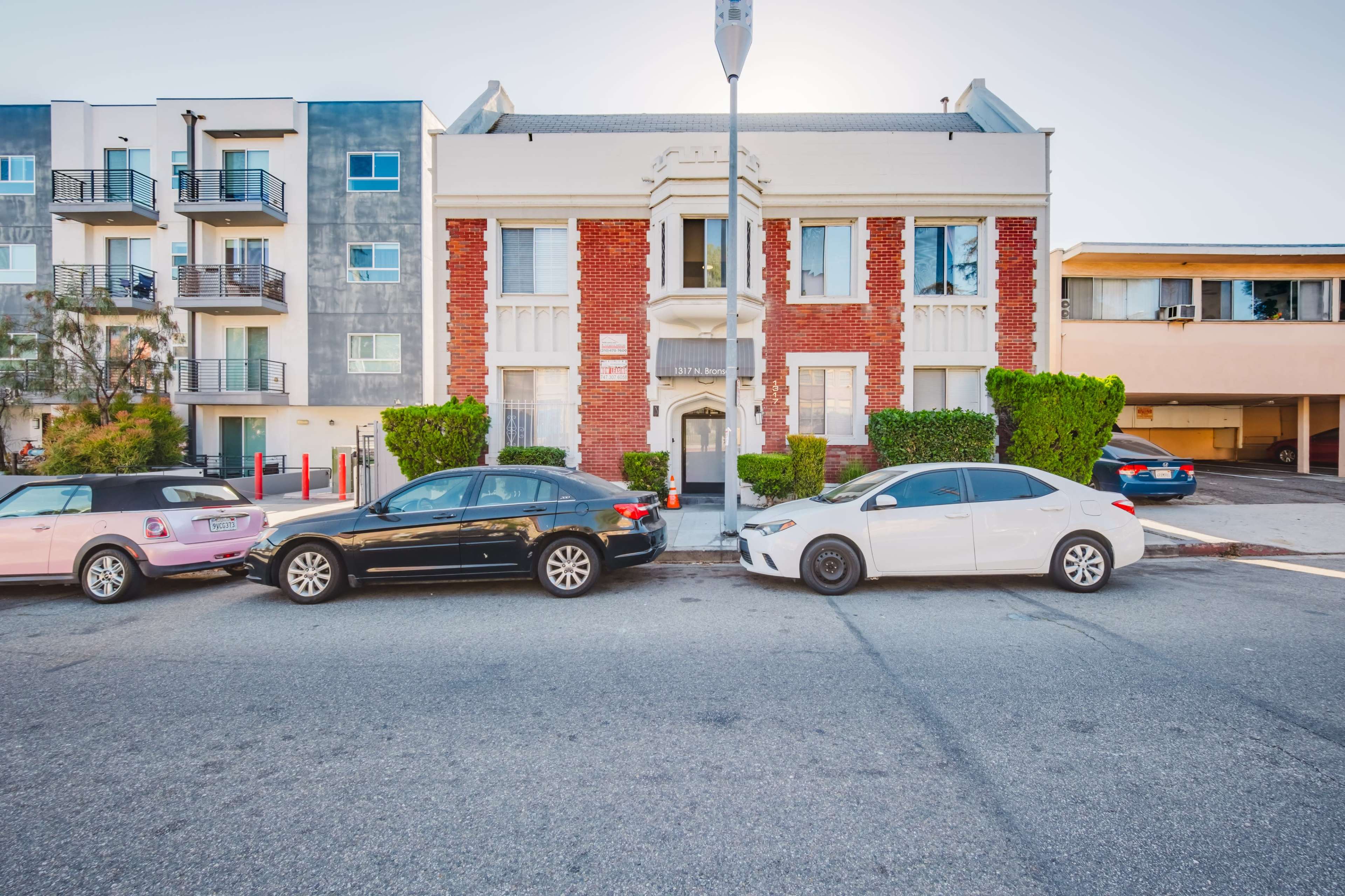 A red brick building with white trim and a sign is flanked by two modern apartment complexes, with several parked cars in front.