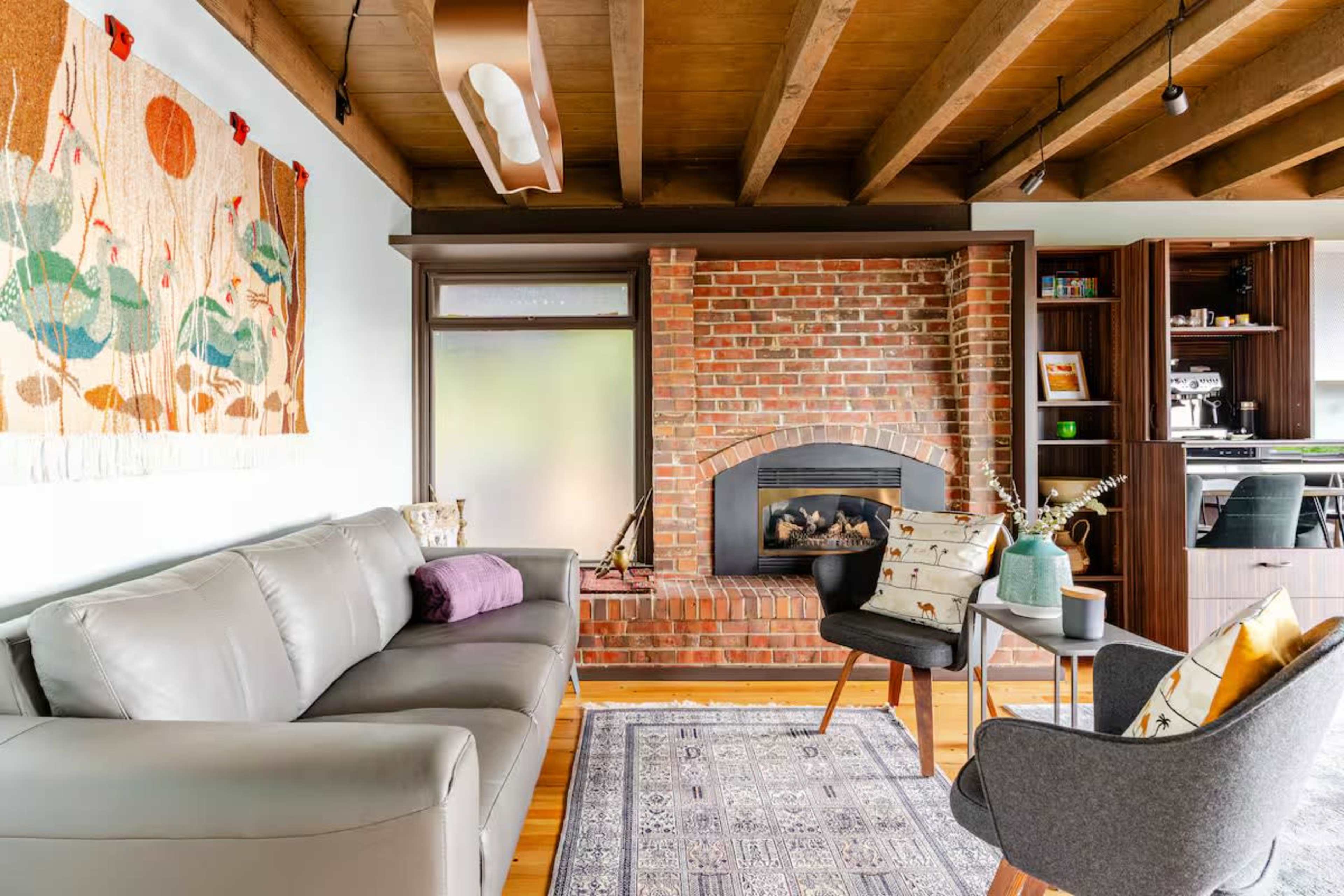 The image shows a modern living room featuring a gray sofa, a brick fireplace, and a tapestry hanging on the wall, with a patterned area rug and stylish chairs arranged around a coffee table.