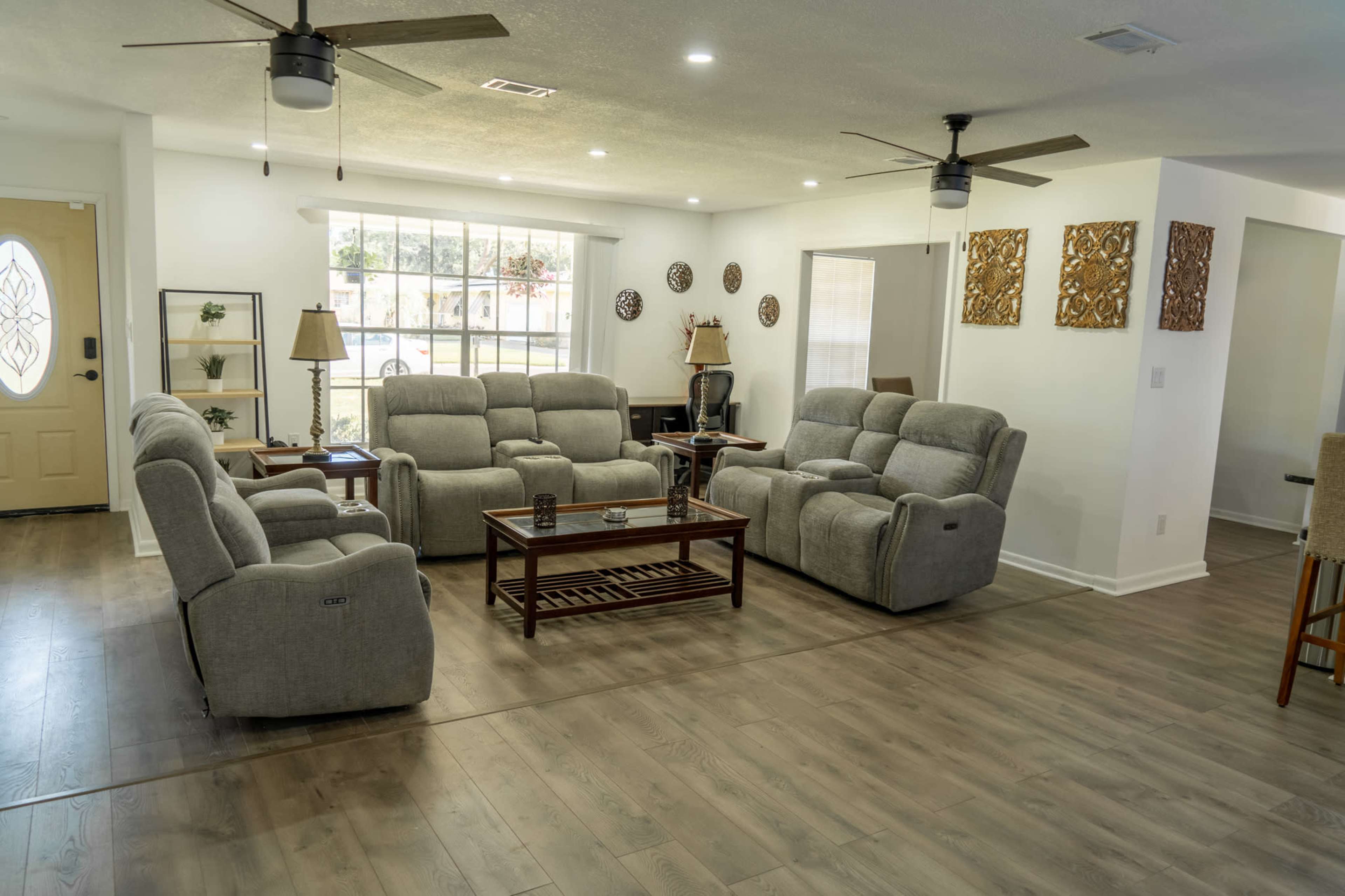The room features a spacious layout with gray reclined sofas arranged around a wooden coffee table, set in front of a large window and a light-colored front door.
