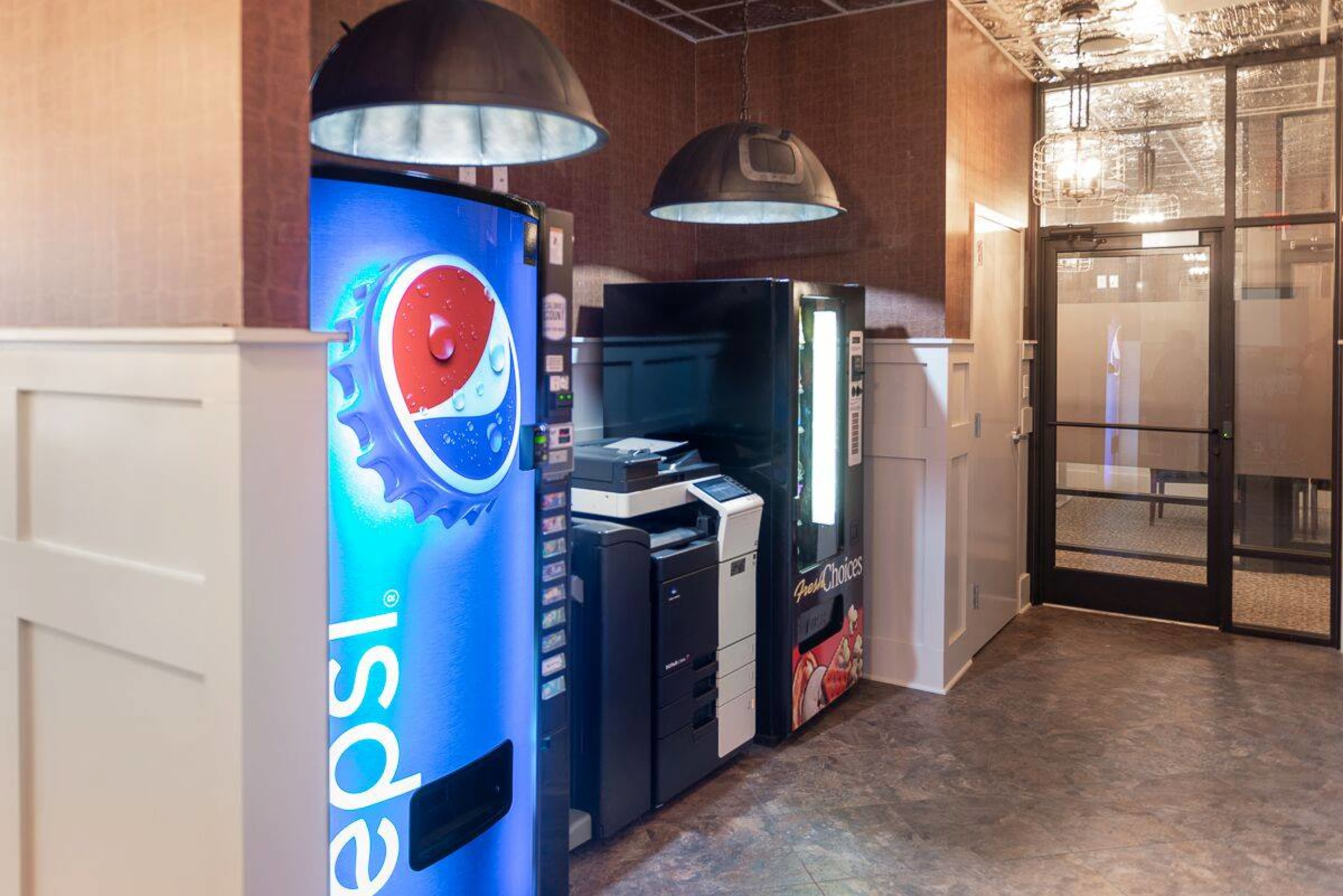 The image shows a vending area featuring a Pepsi vending machine, a printer, and a snack vending machine, with an adjacent glass door leading to another room.
