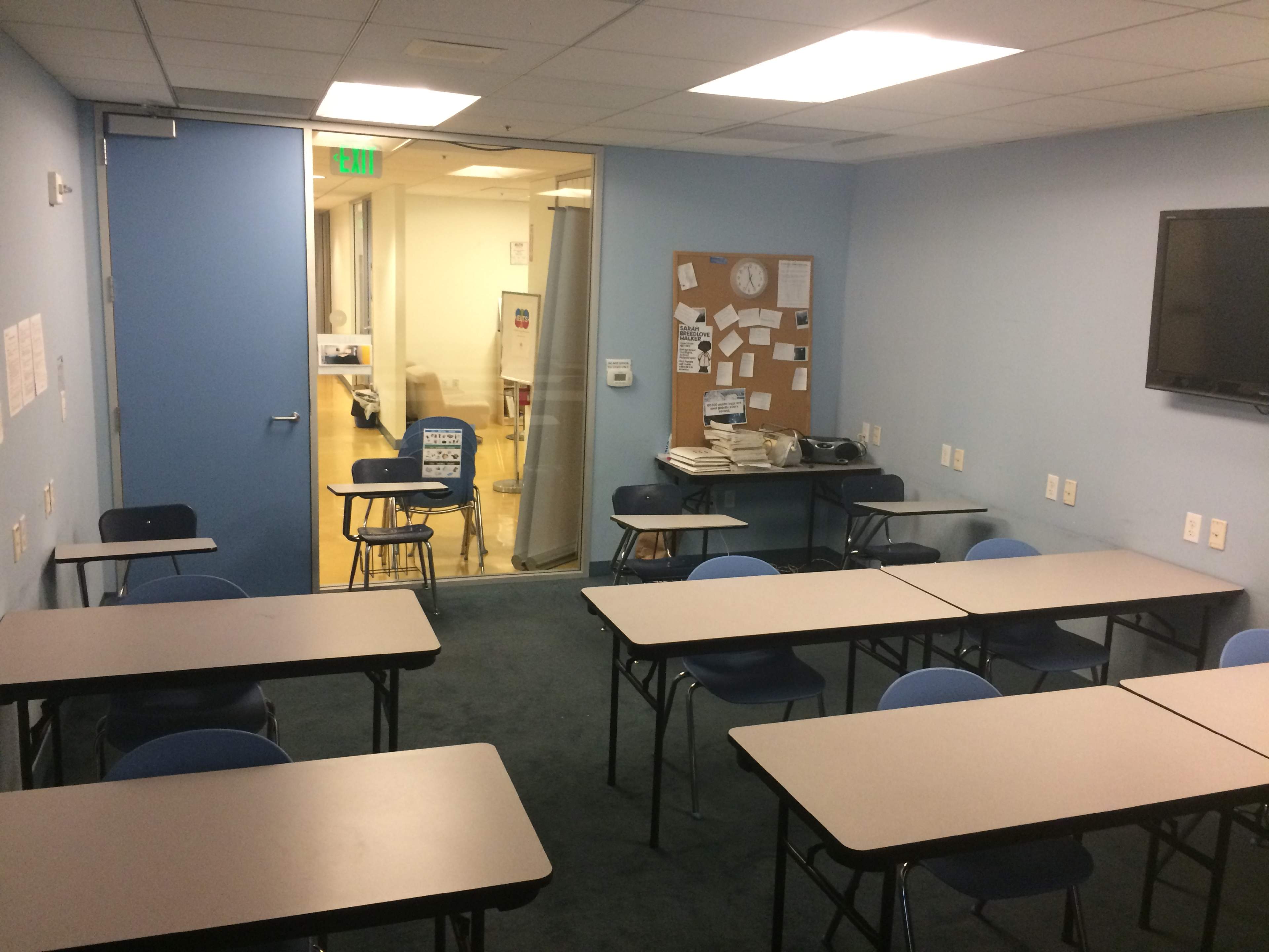 A small classroom with several desks and chairs arranged in rows, a bulletin board on the wall, and a door leading to another area.