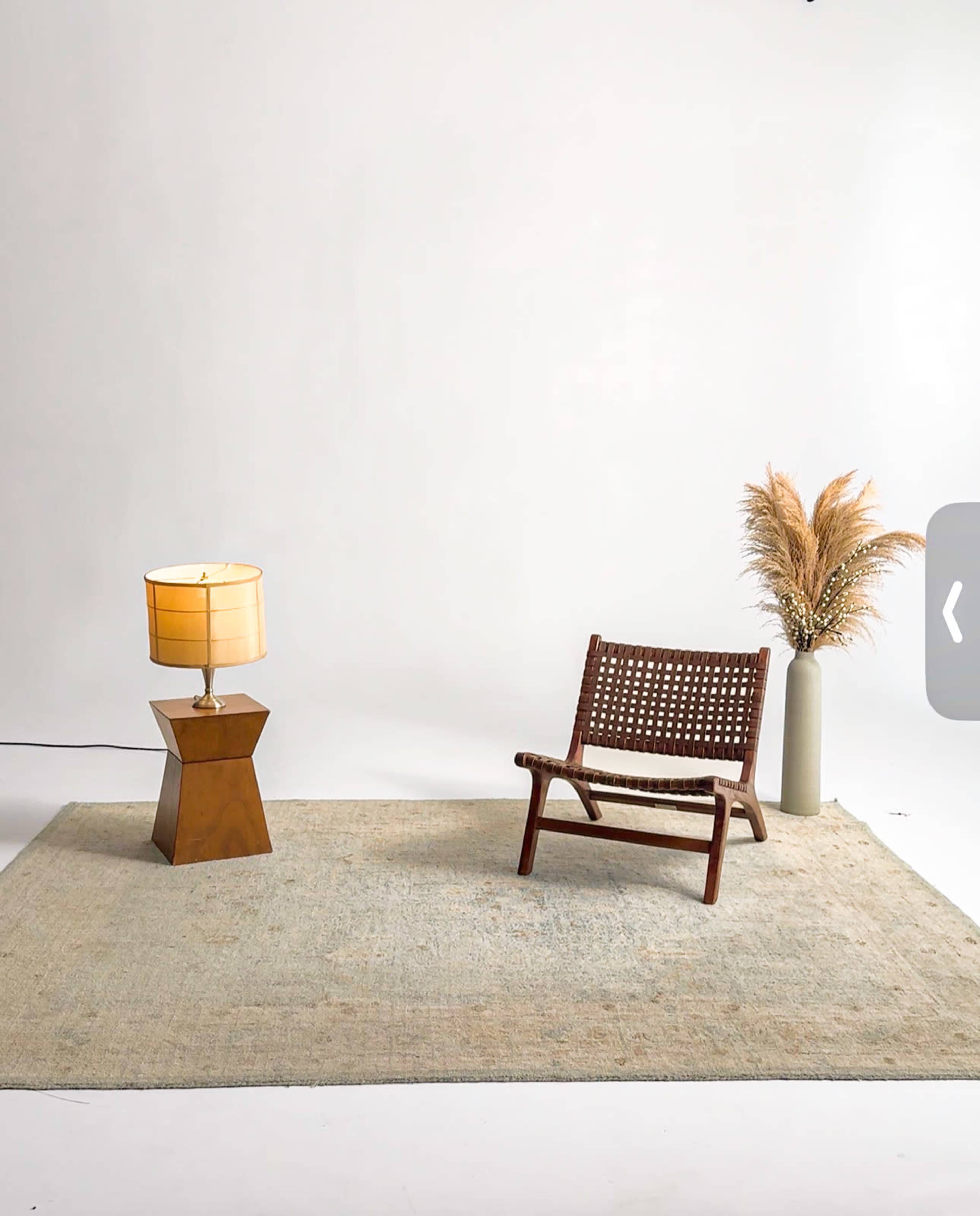 The image features a minimalist interior scene with a wooden chair, a lamp with a fabric shade, a textured rug, and a decorative vase containing dried plants.