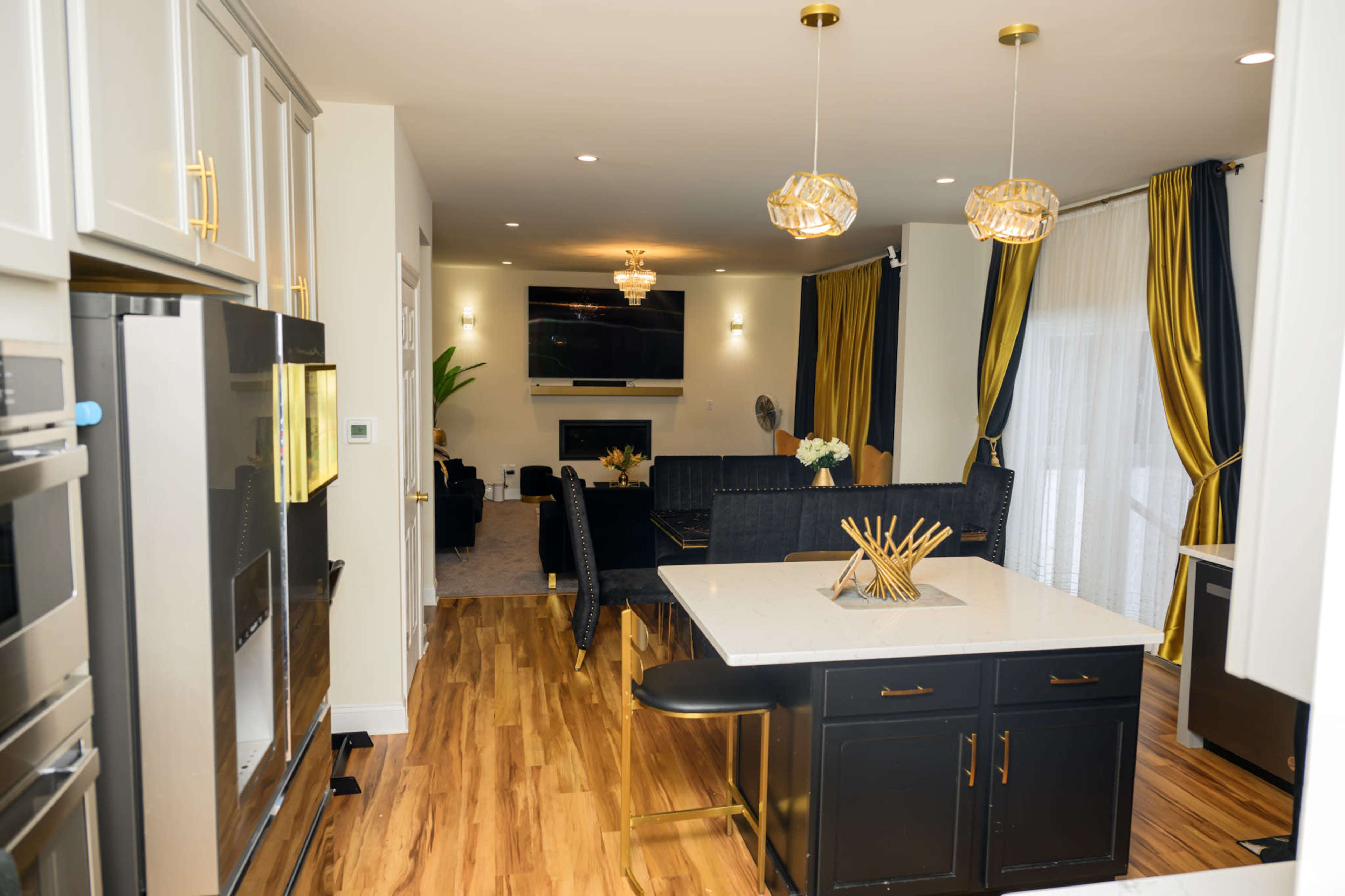 The image features a modern kitchen with a central island, equipped with sleek appliances, and opens into a living area adorned with decorative curtains and a wall-mounted television.