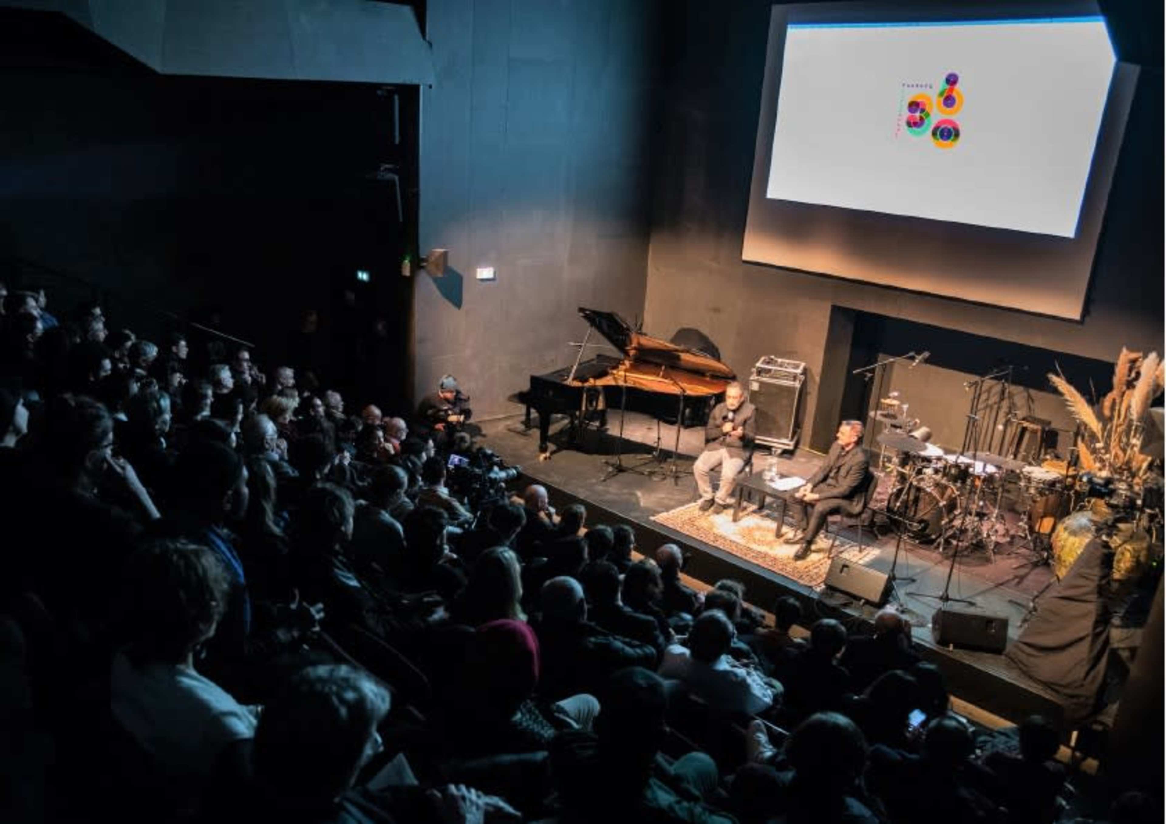A large audience watches a discussion on stage with a piano and a band setup in the background.
