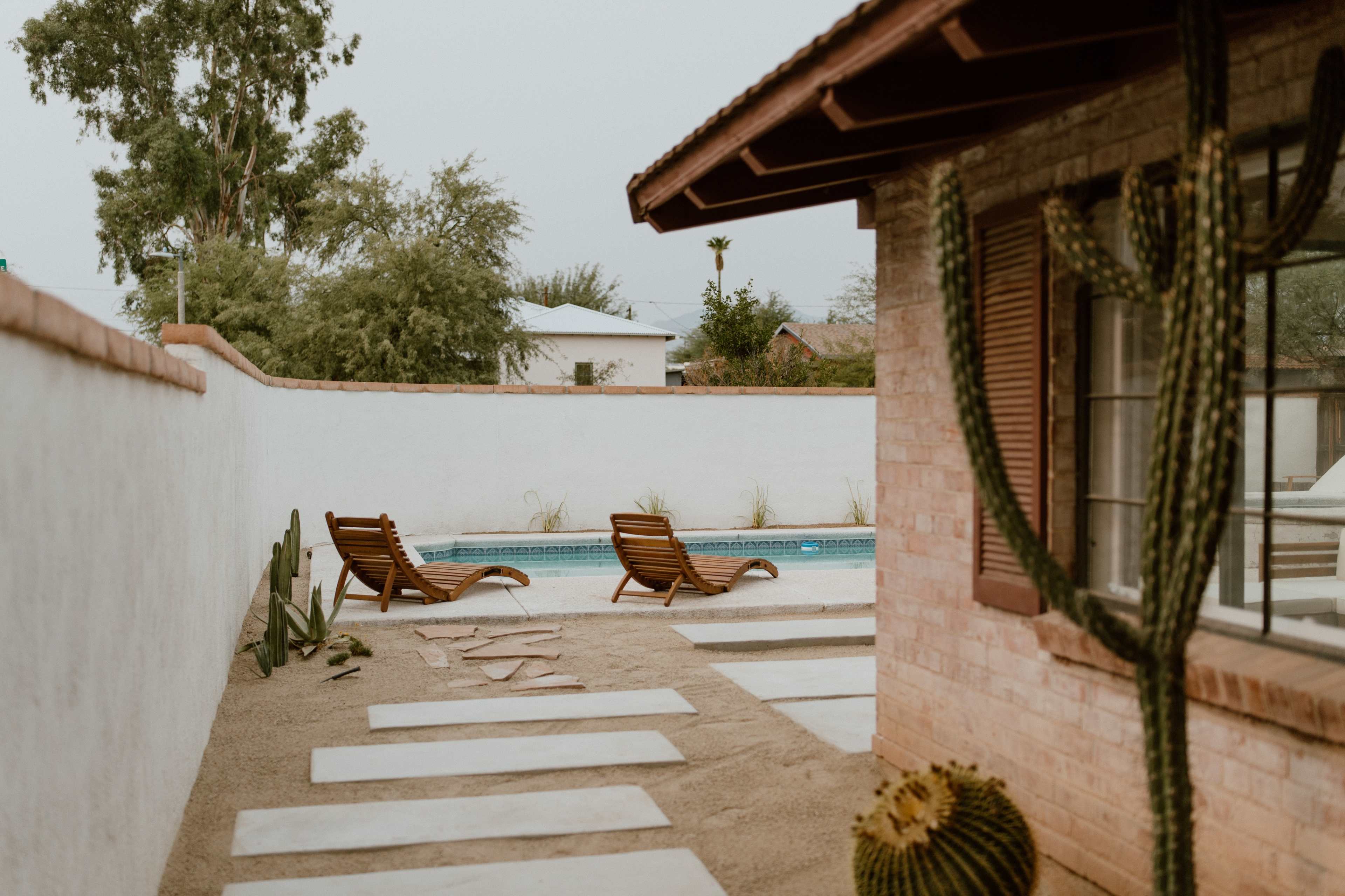 A view of a pool area featuring two lounge chairs, surrounded by a sandy landscape with cacti and stepping stones.
