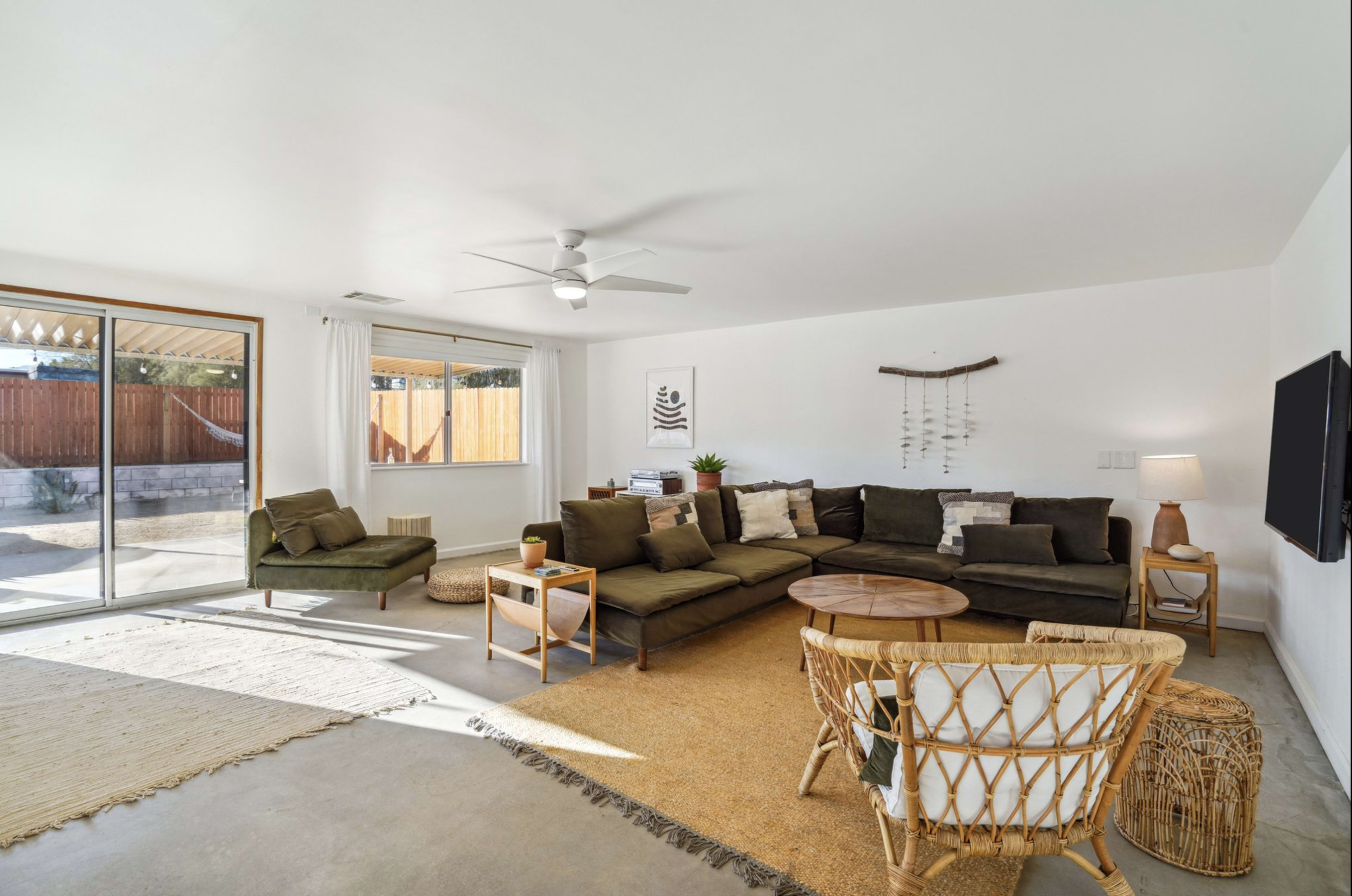 The living room features a spacious sectional sofa, a round coffee table, and large windows that provide natural light, with concrete flooring and minimal decor.