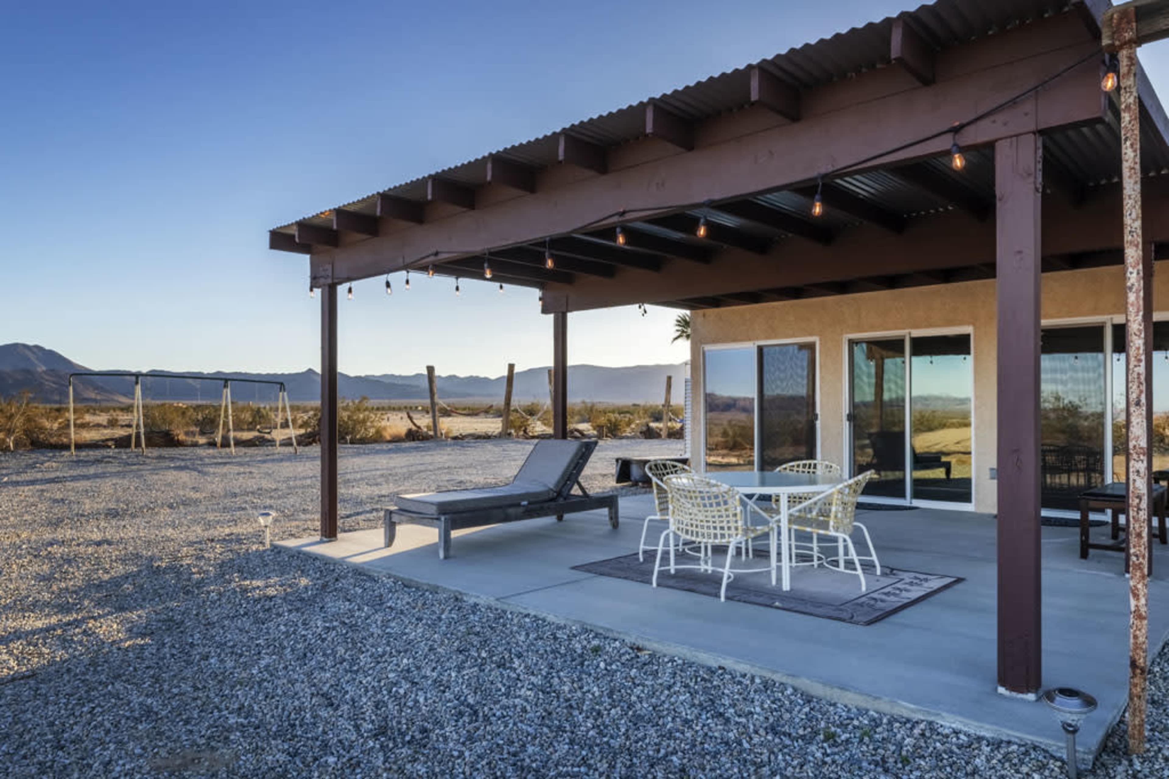 A patio area of a house with a table and chairs, surrounded by gravel and mountain scenery in the background.