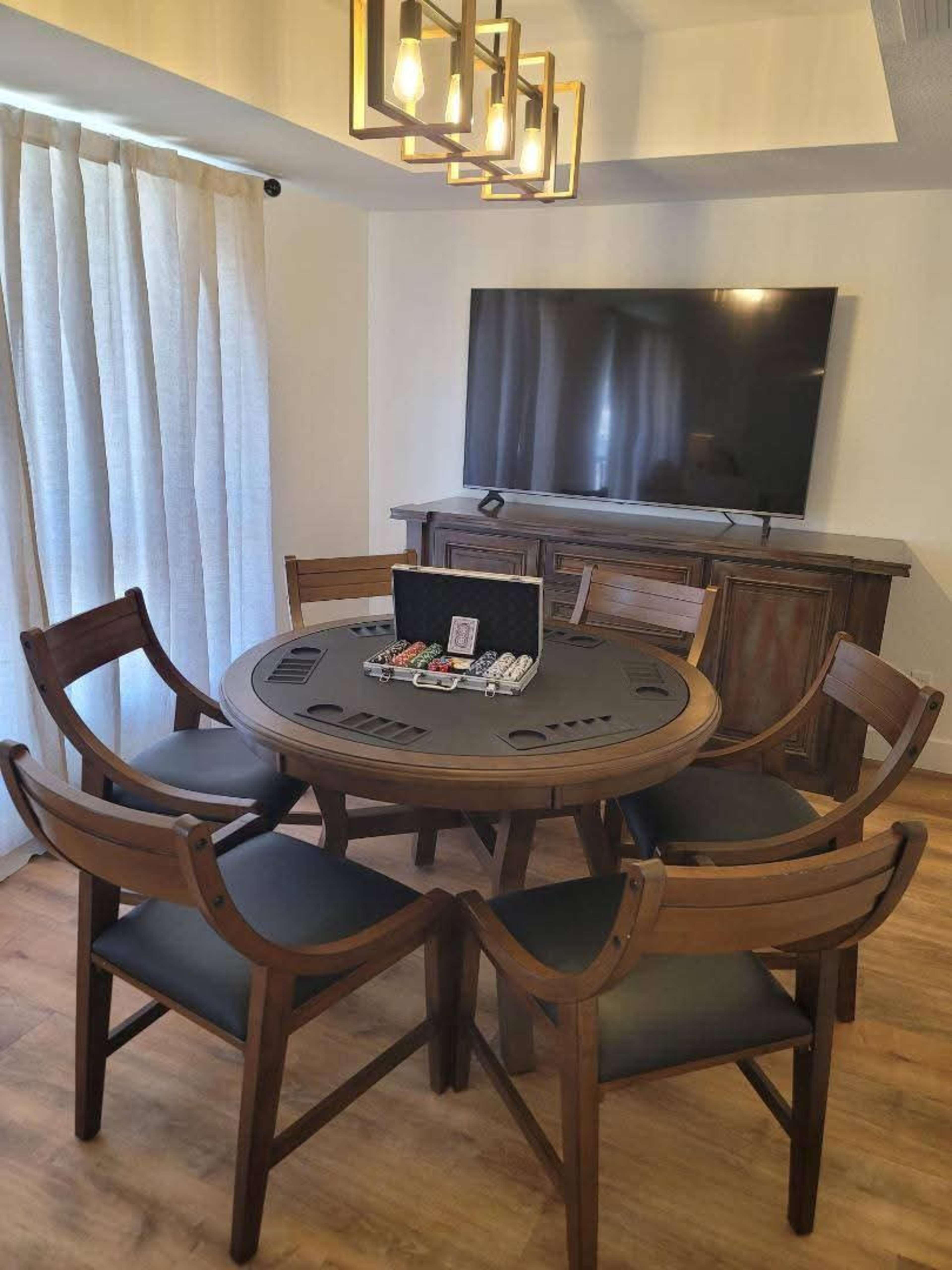 A round wooden dining table with six chairs is set in a room featuring a television mounted on the wall and a curtain covering a window.