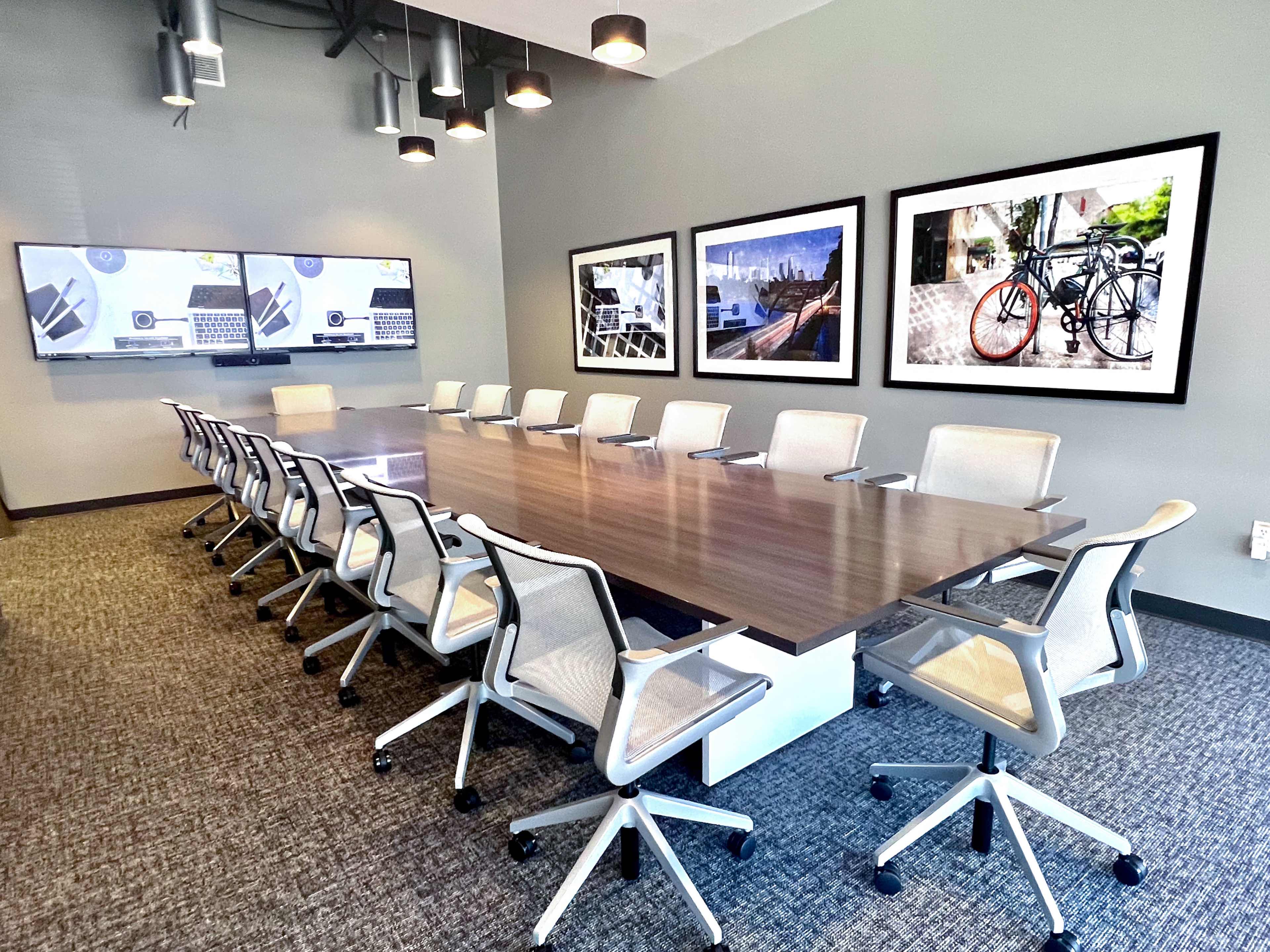 A modern conference room features a long table surrounded by ergonomic chairs and two large screens mounted on the walls, with framed photographs of cityscapes and bicycles.