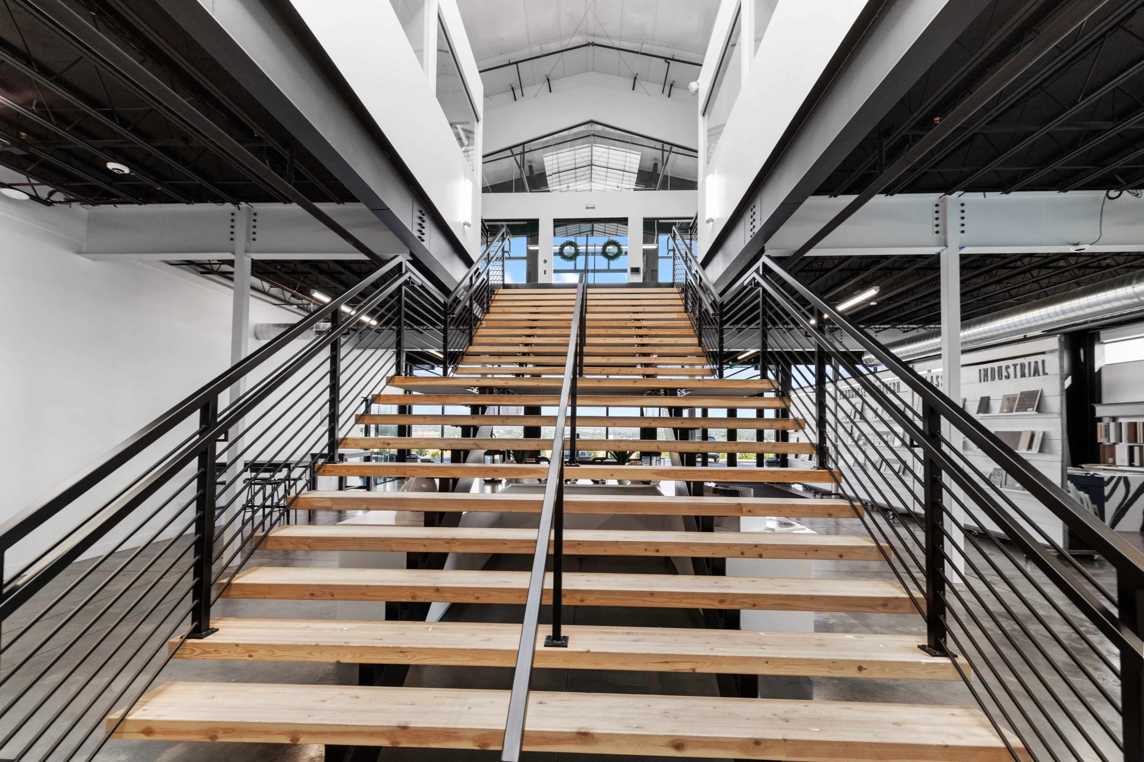 The image shows a modern wooden staircase with black railings leading to a bright, open space with large glass doors and high ceilings.