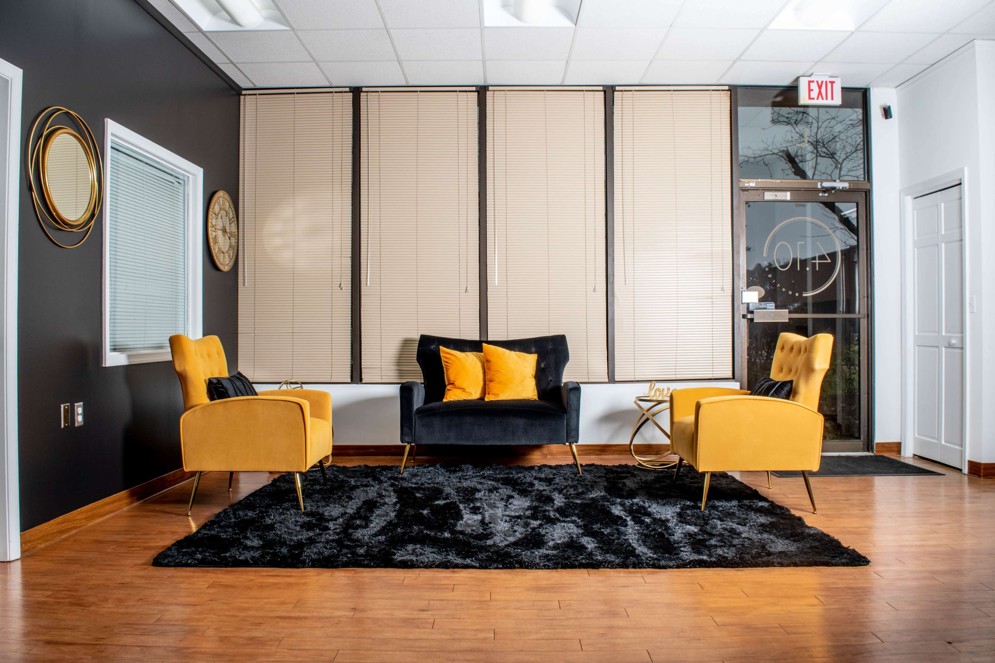 A modern waiting room featuring two yellow chairs, a black sofa, a coffee table, and a large black rug, with large windows covered by blinds.
