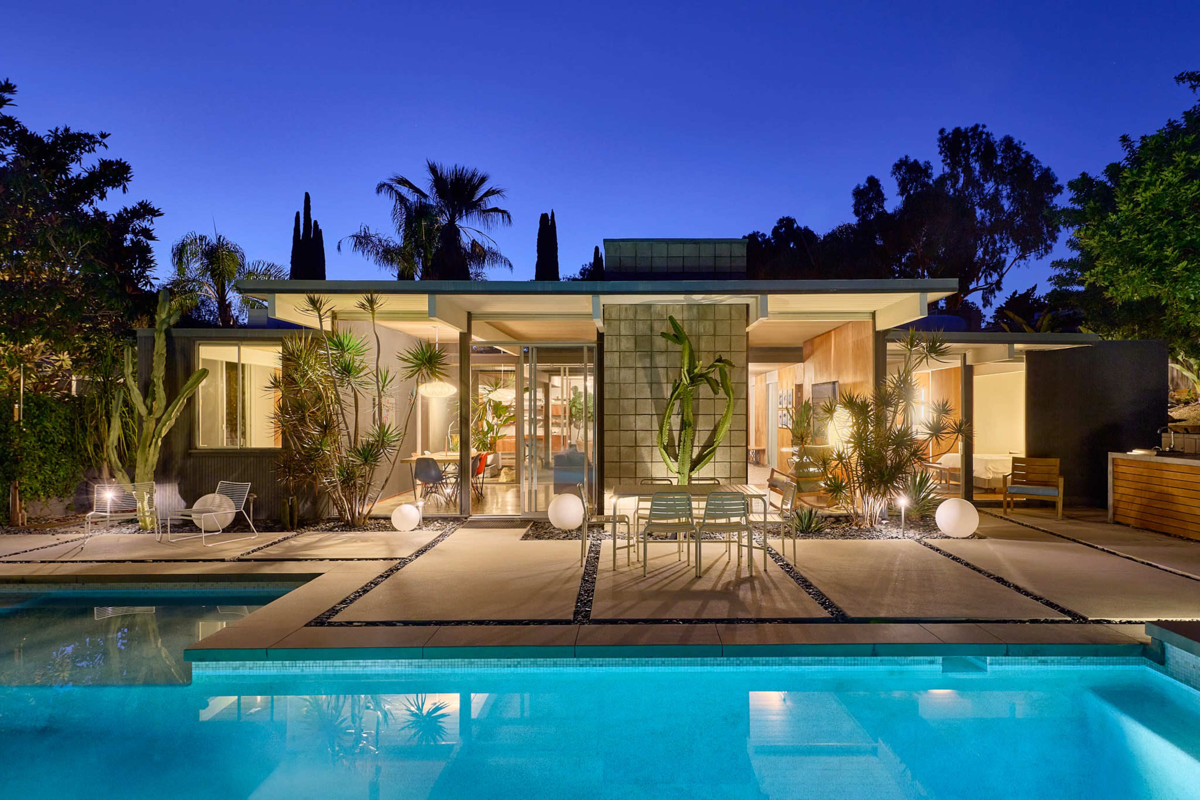 A modern house with large glass windows and a swimming pool is illuminated by evening light, surrounded by tropical plants.